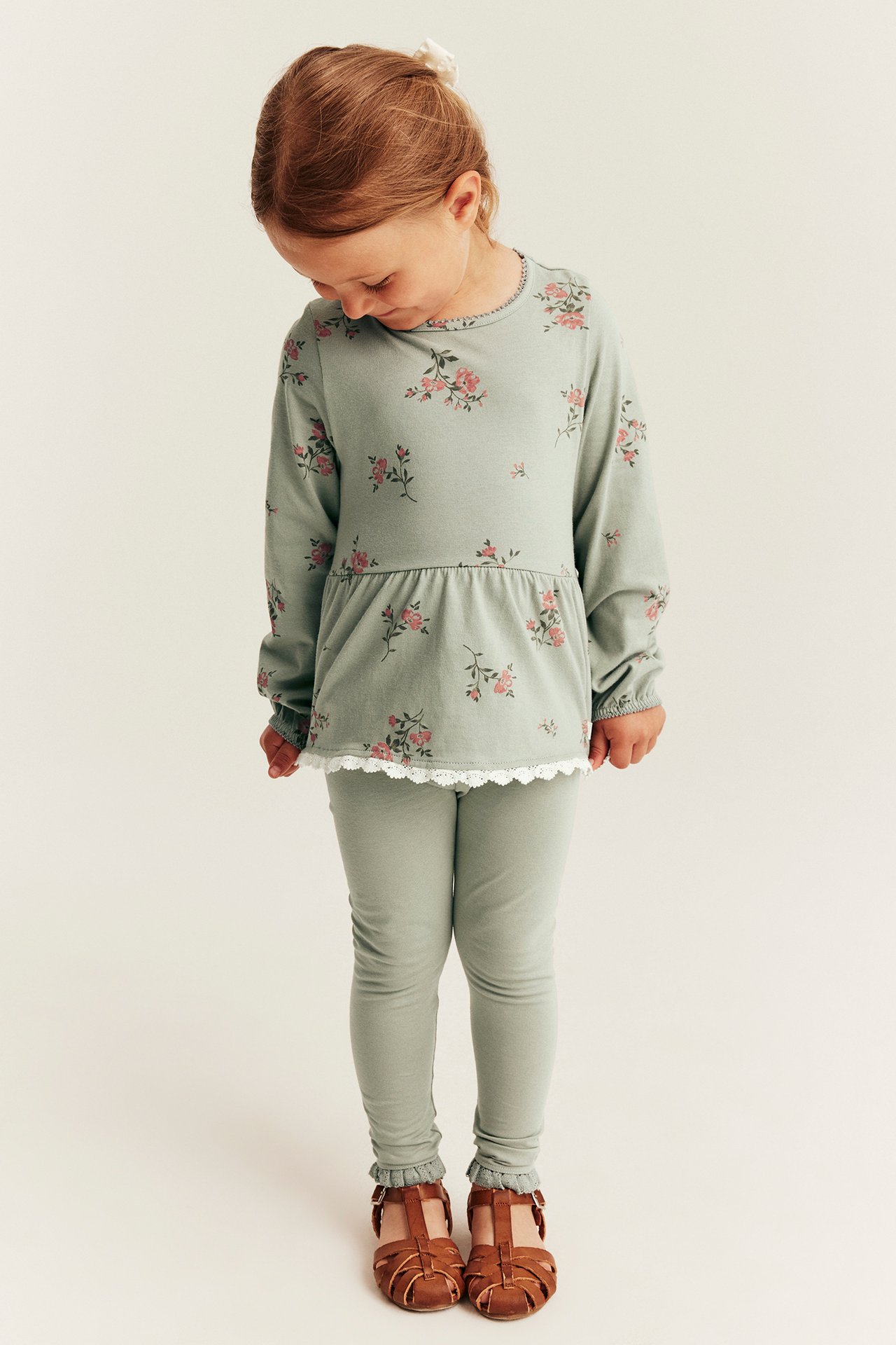 Forfra: Et barn i en grønn blomstret langermet peplumtopp med blondekant, grønne leggings og brune sandaler.