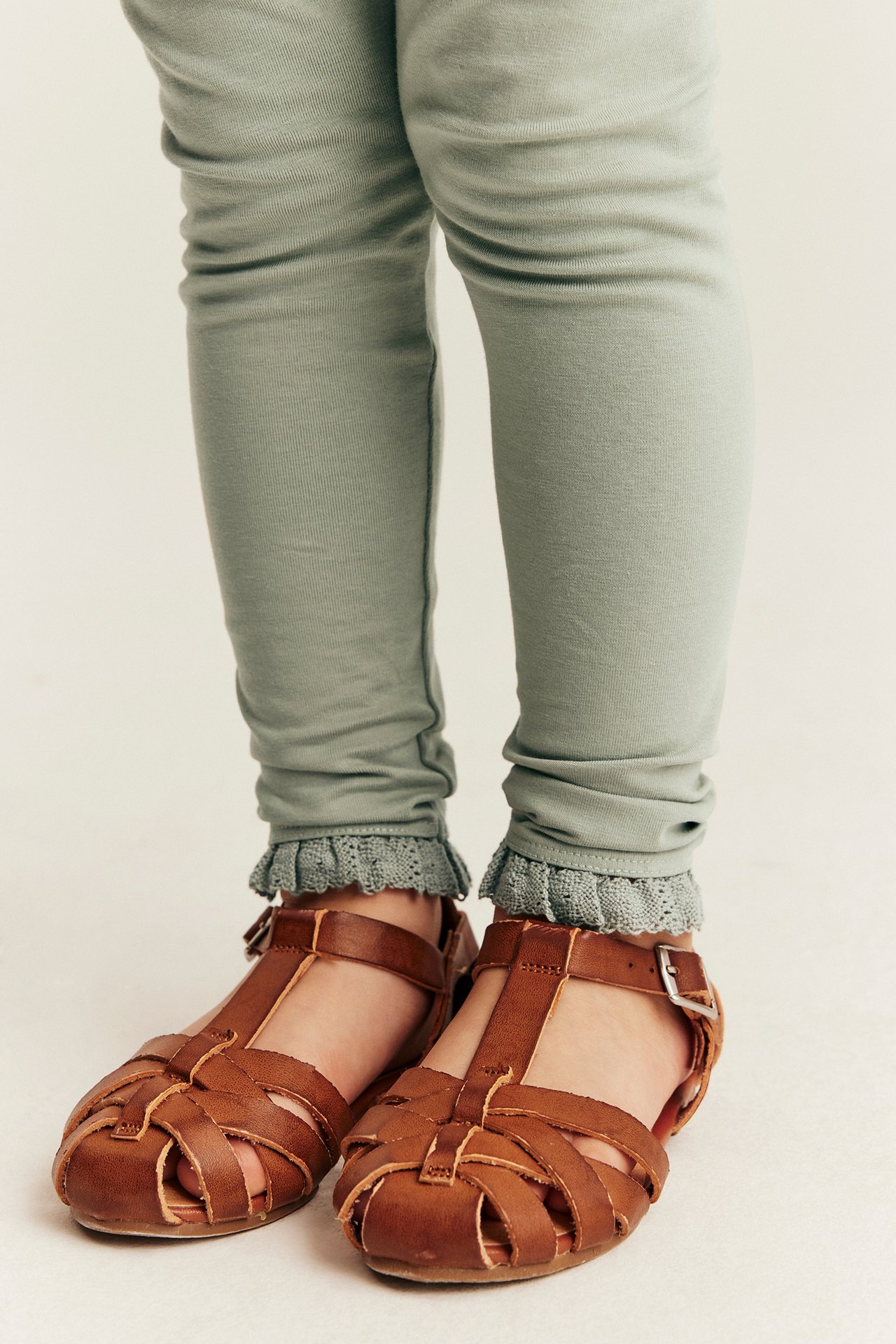 Nærbilde, frontvisning av et barn i lysegrønne leggings med gråblå blondekant og brune skinnsandaler.