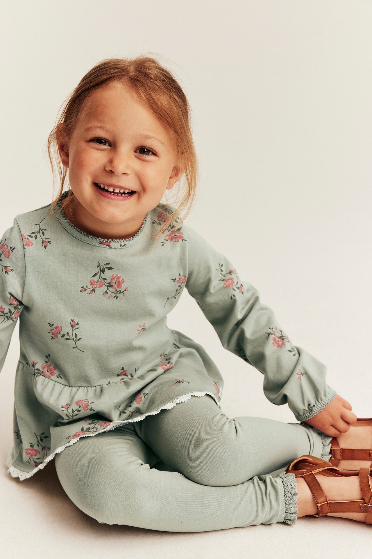 Frontbilde: Barn i grønn blomstret peplumtopp, lysegrønne leggings og brune sandaler.