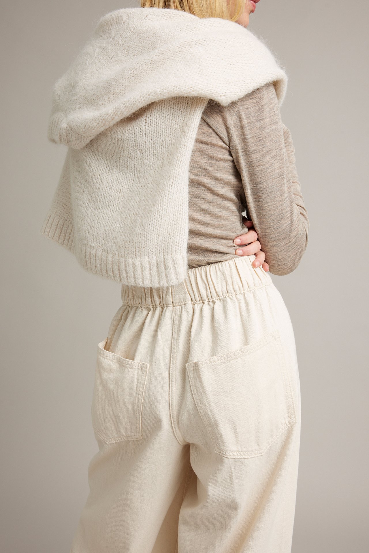 Bakifrån kvinna i benvit stickad huvväst, beige topp och ljusbeige byxor med resår.