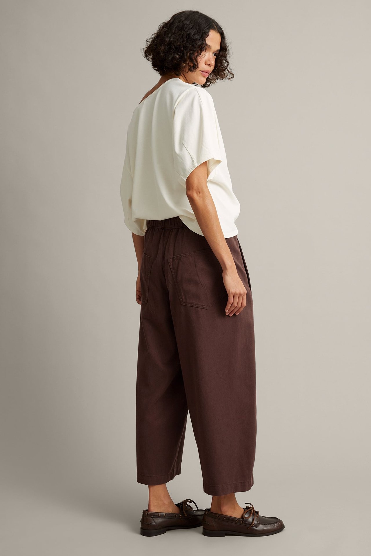 Kvinnelig modell sideprofil i mørkebrune vide bukser, kremfarget kortermet topp og mørkebrune loafers.