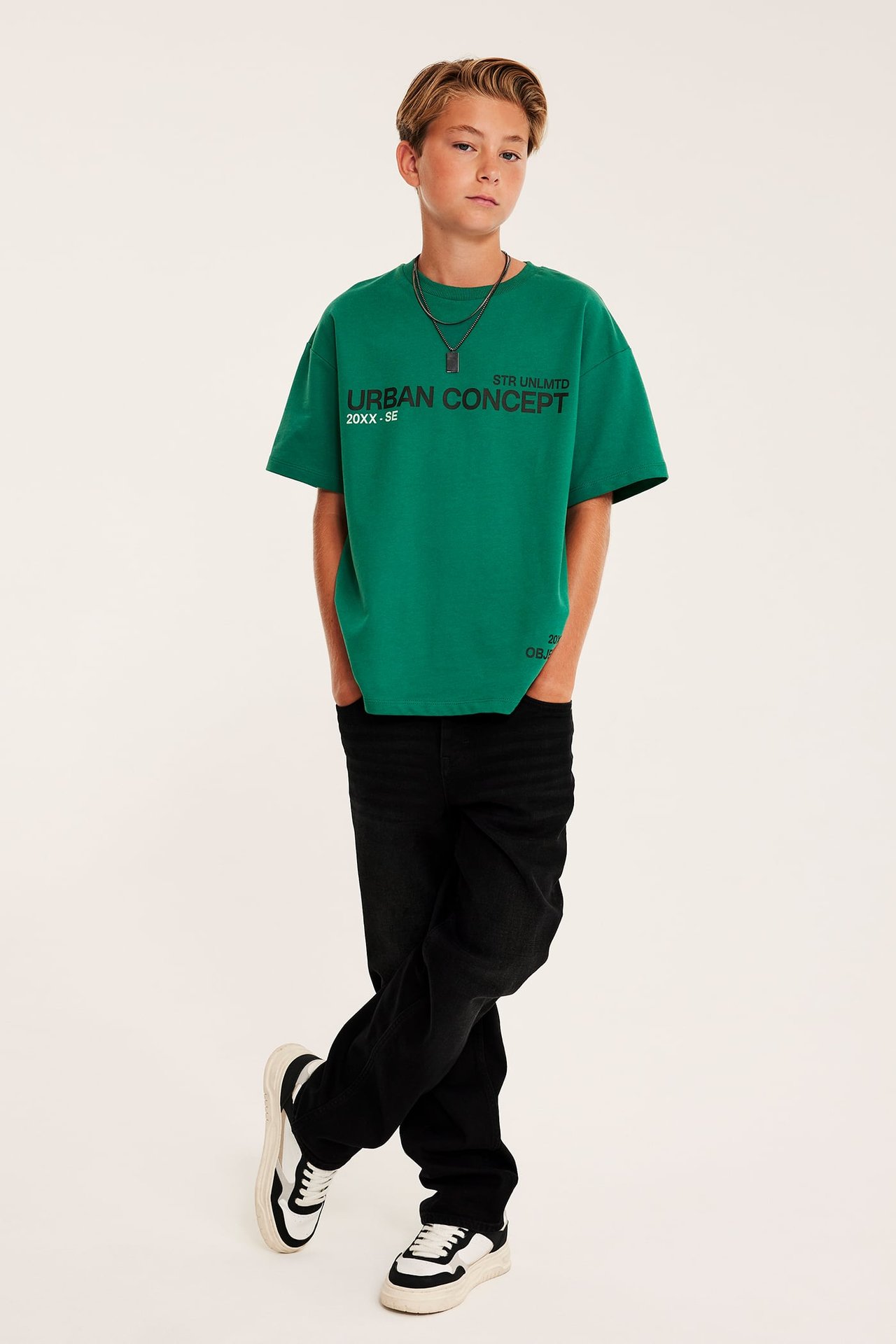 Framifrån: barn i grön URBAN CONCEPT t-shirt, svarta jeans, sneakers och silverhalsband.