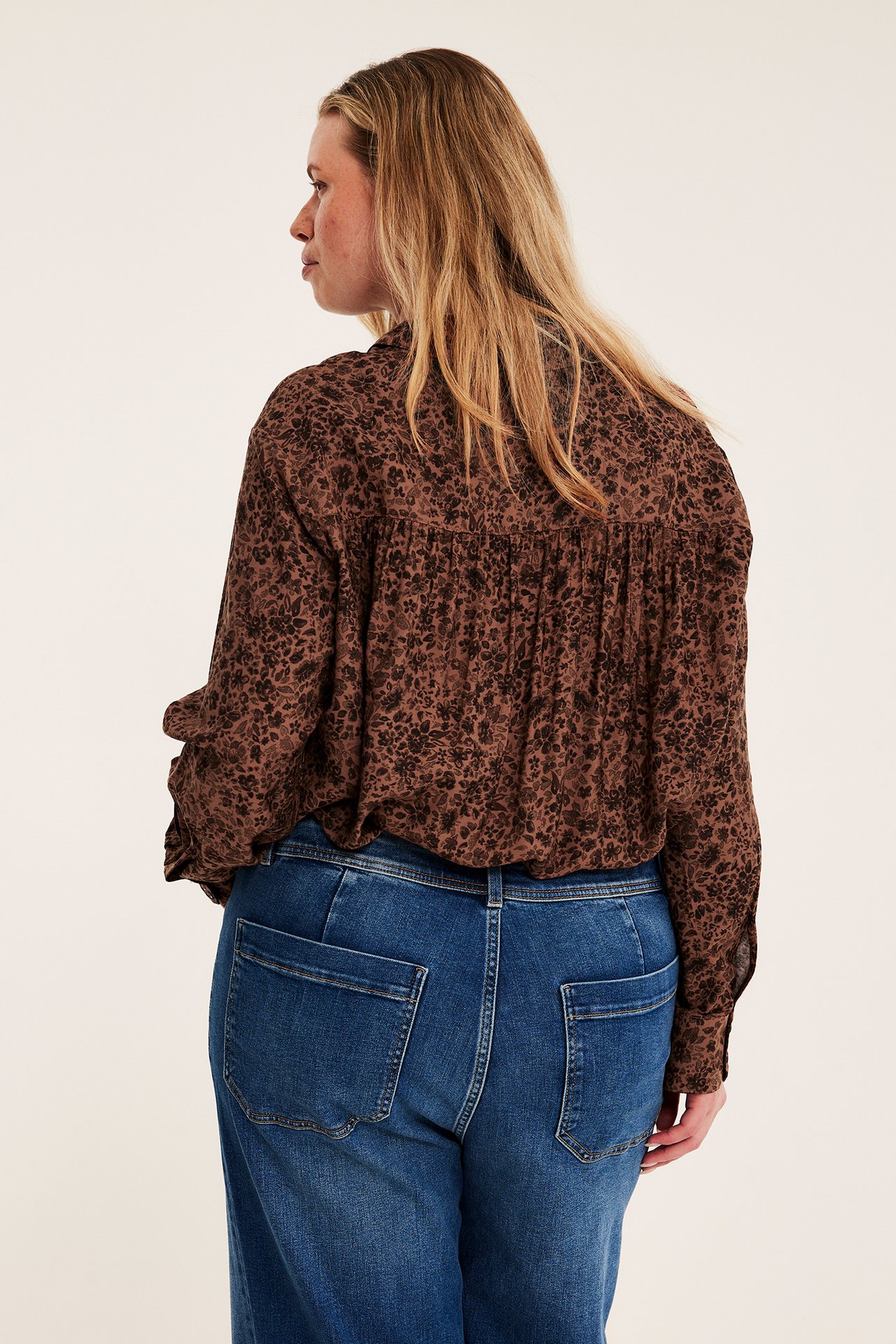 Ryggbilde av kvinnelig modell iført brunmønstret løstsittende skjorte og blå jeans.