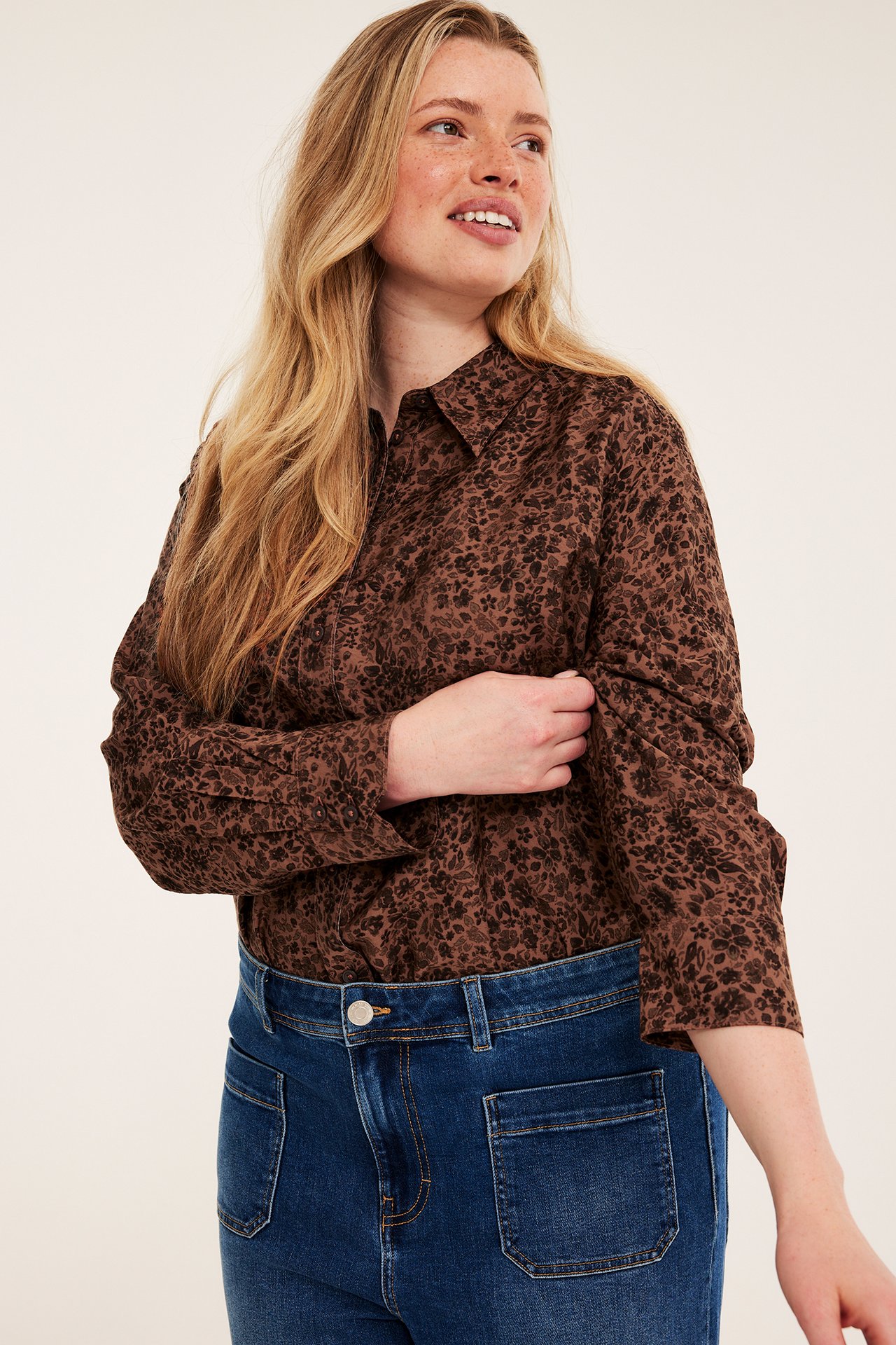 Forfra-side visning av kvinne i brun mønstret skjorte, rullende ermet, og blå jeans.