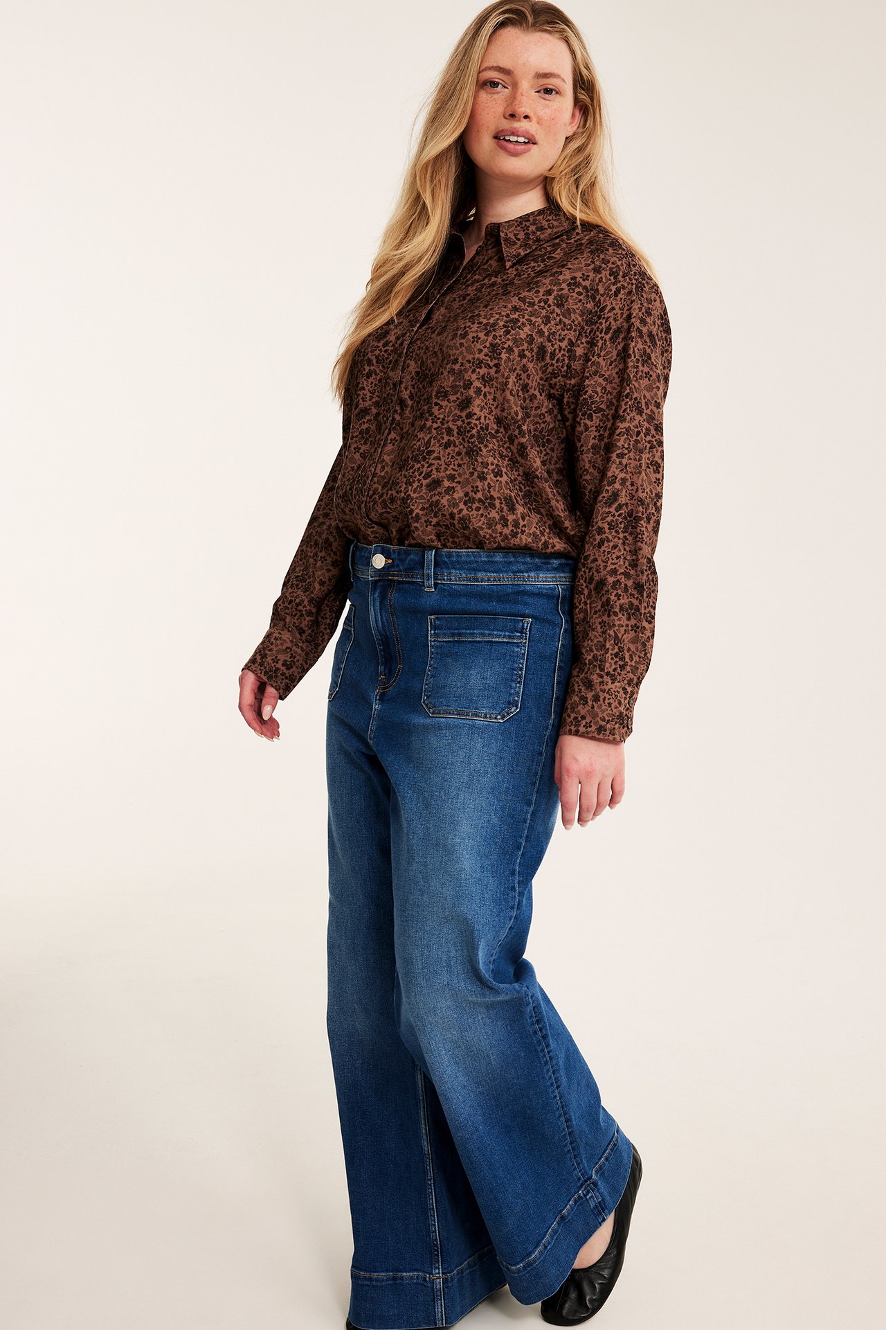 Frontbilde av kvinne i brun blomstret langermet skjorte, blå vide jeans med frontlommer og svarte ballerinasko.