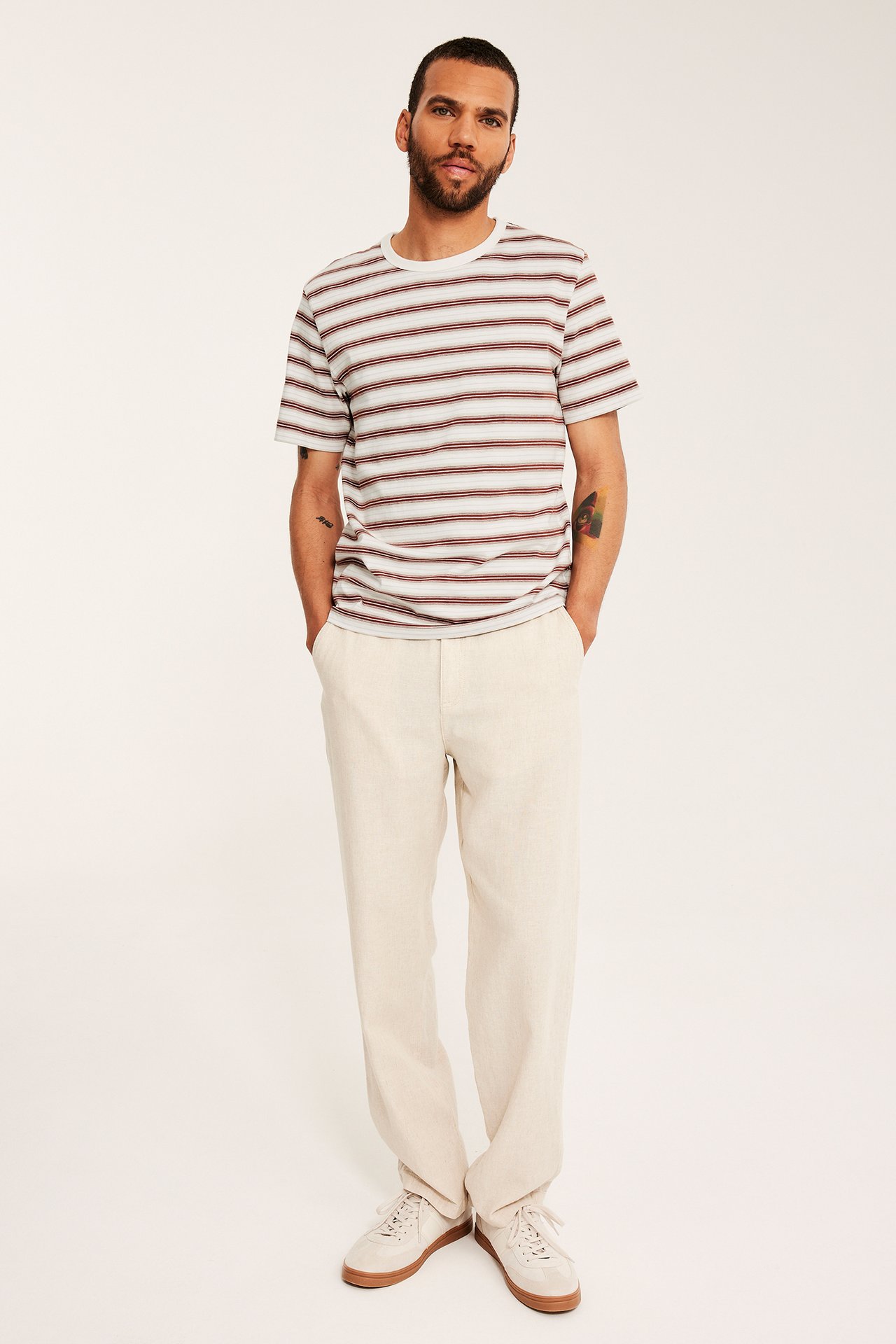 Edestä: mies yllään valkoinen regular fit t-paita ruskeilla raidoilla, vaaleanbeiget housut ja kermaiset lenkkarit.