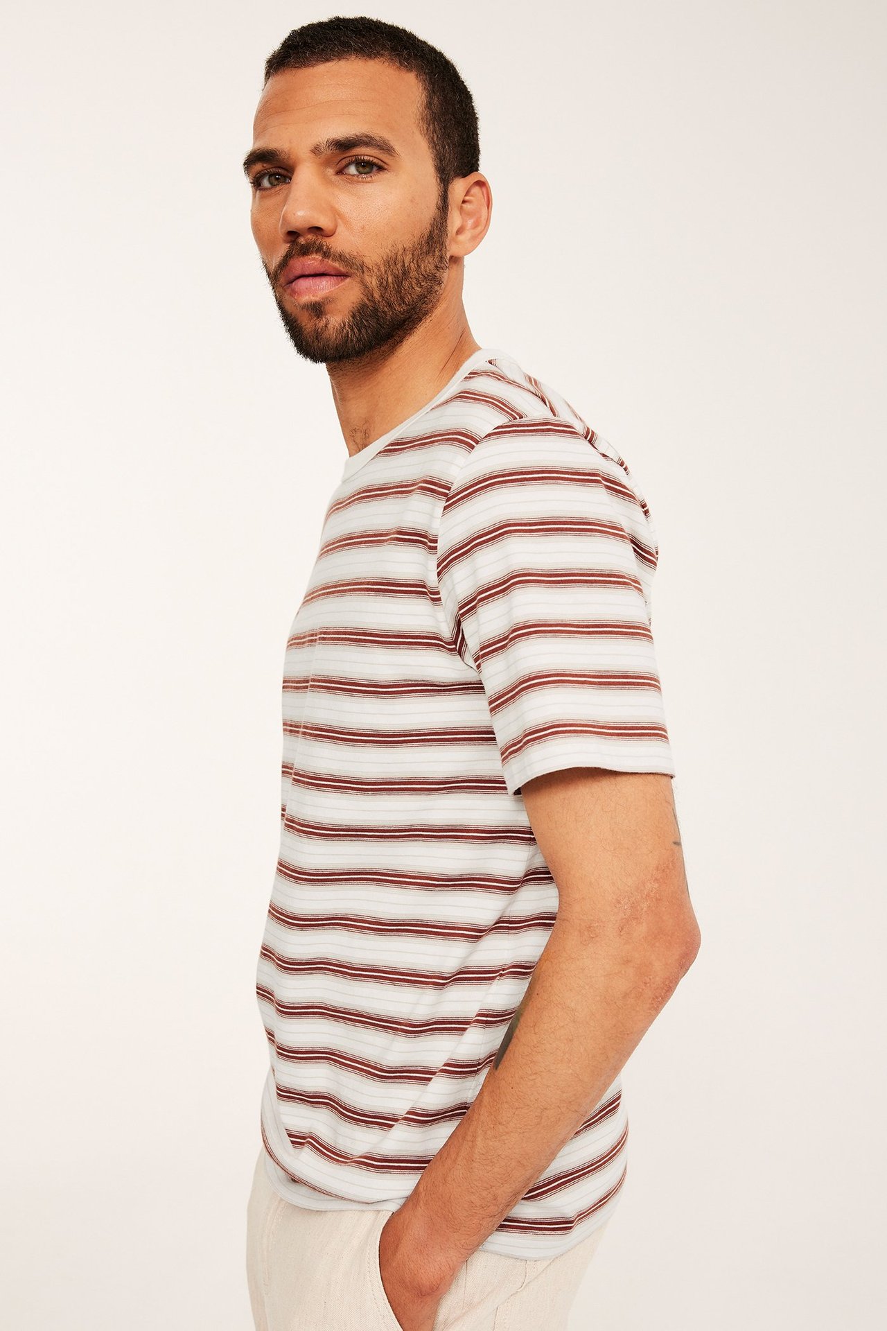 Mies sivukuvassa pukeutuneena ruskea-valkoraidalliseen puuvilla-T-paitaan ja vaaleanbeigeihin housuihin.
