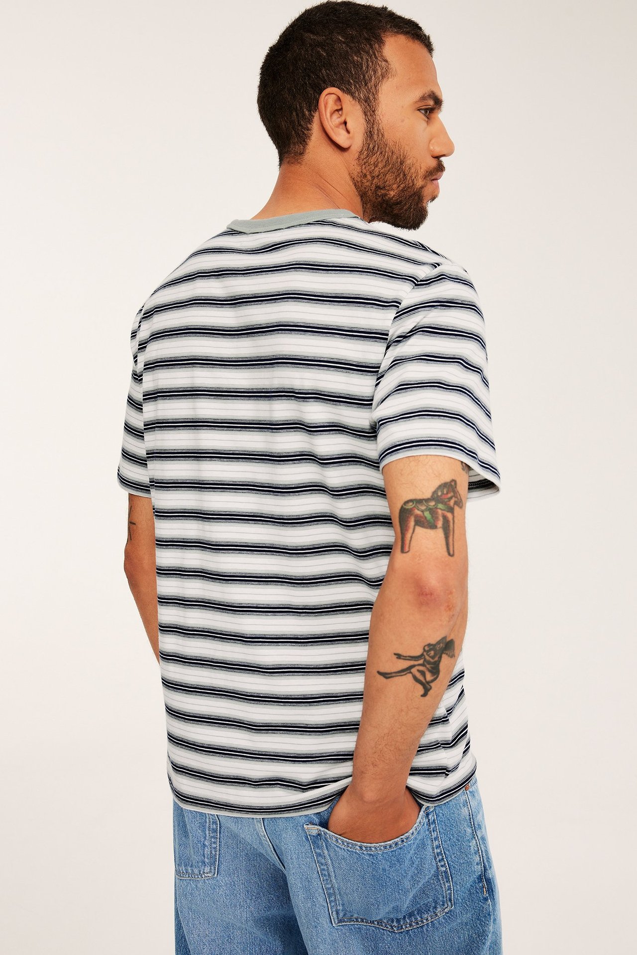 Mies tummansinisessä ja valkoisessa raidallisessa puuvilla-t-paidassa ja vaaleansinisissä farkuissa, takaa sivulta.