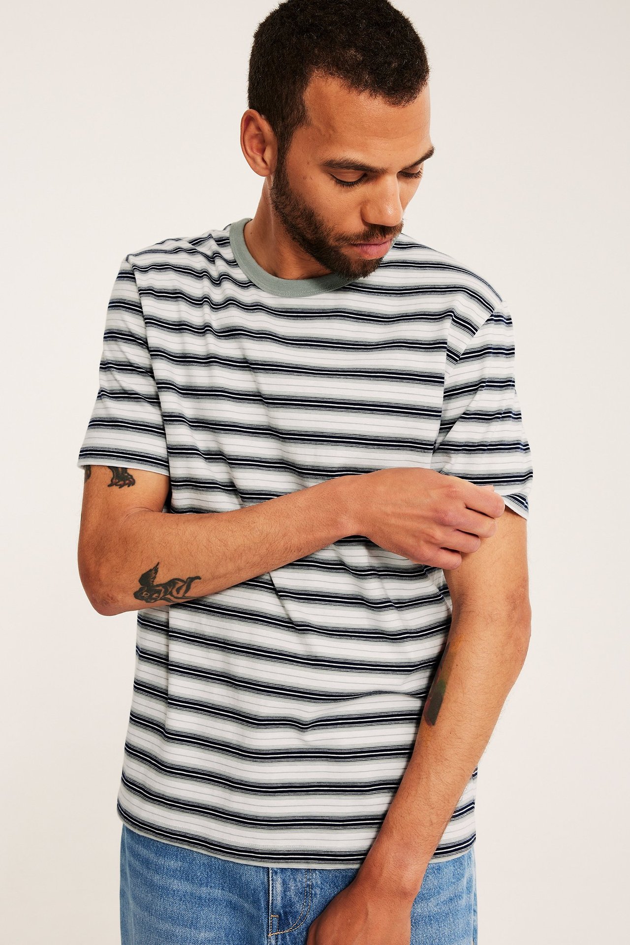 Mies yllään valkoinen, harmaa, tummansininen raidallinen puuvilla t-paita ja siniset farkut.