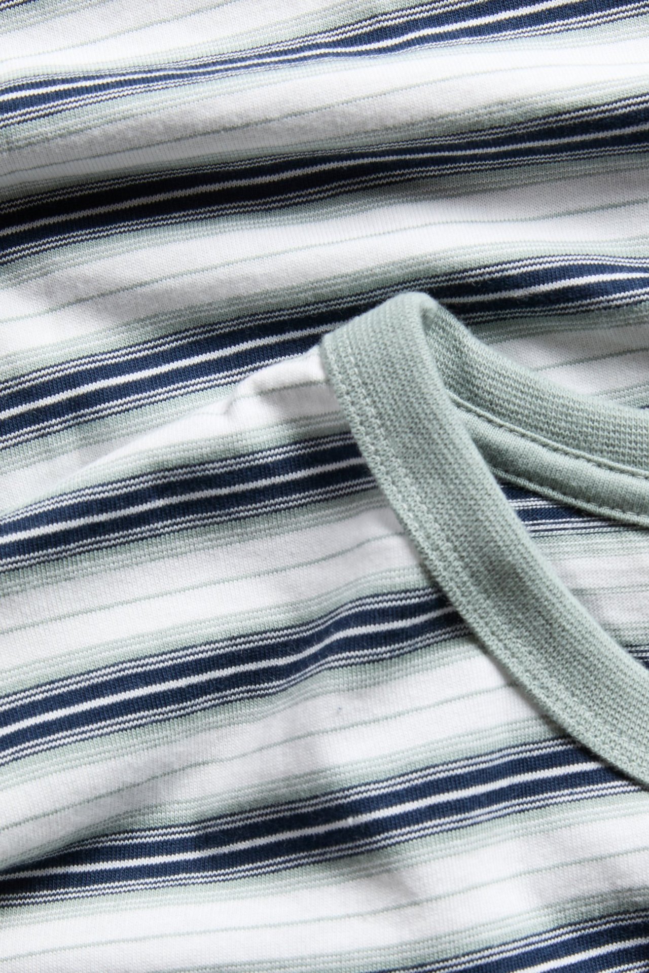 Zbliżenie: Bawełniany T-shirt w paski z niebieskimi, białymi i stonowanymi zielonymi paskami, oraz zielonym prążkowanym dekoltem.