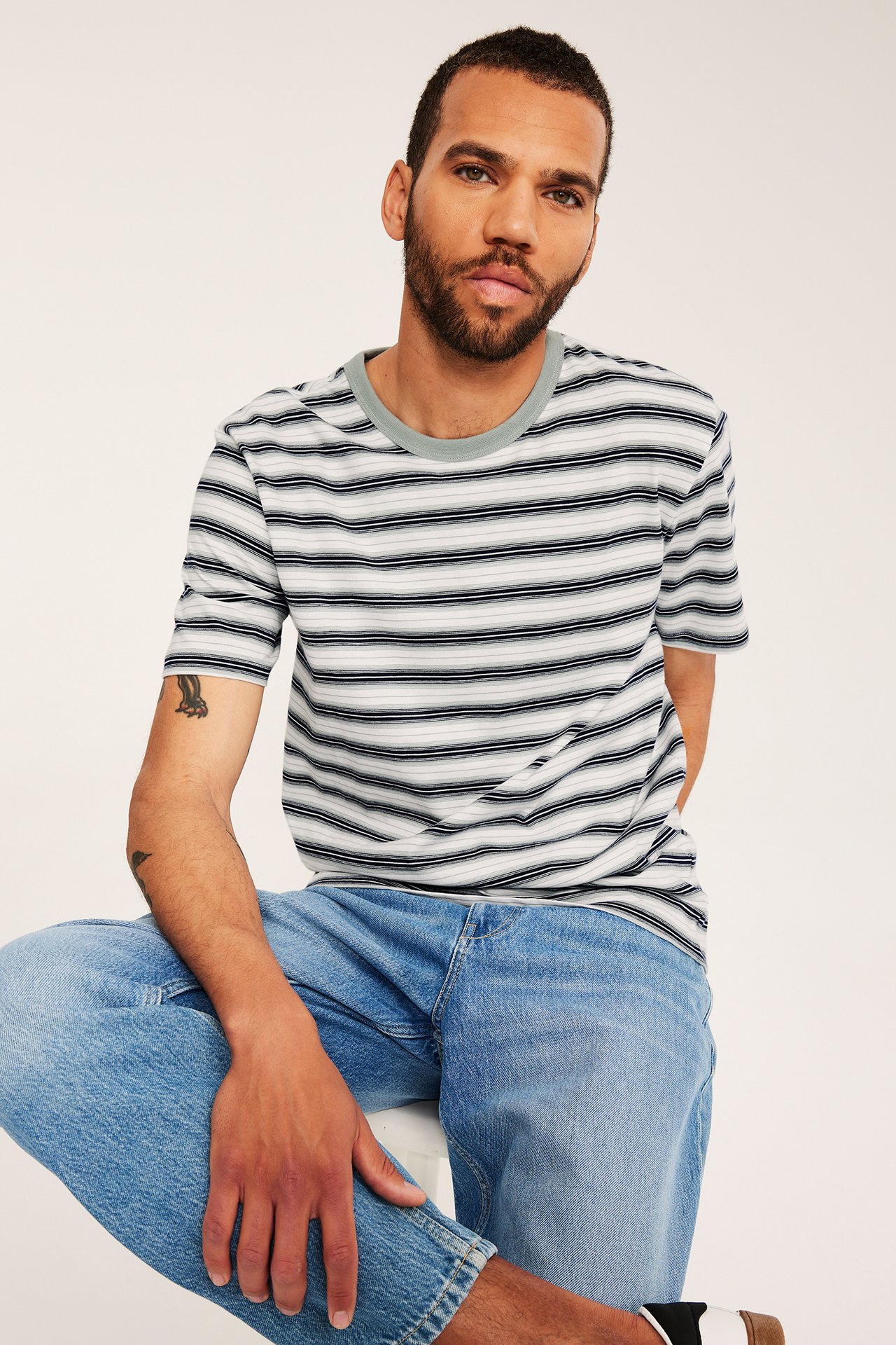 Edestä: Mies raidallisessa puuvilla-T-paidassa, vihreä pyöreä pääntie, ja sinisissä farkuissa.