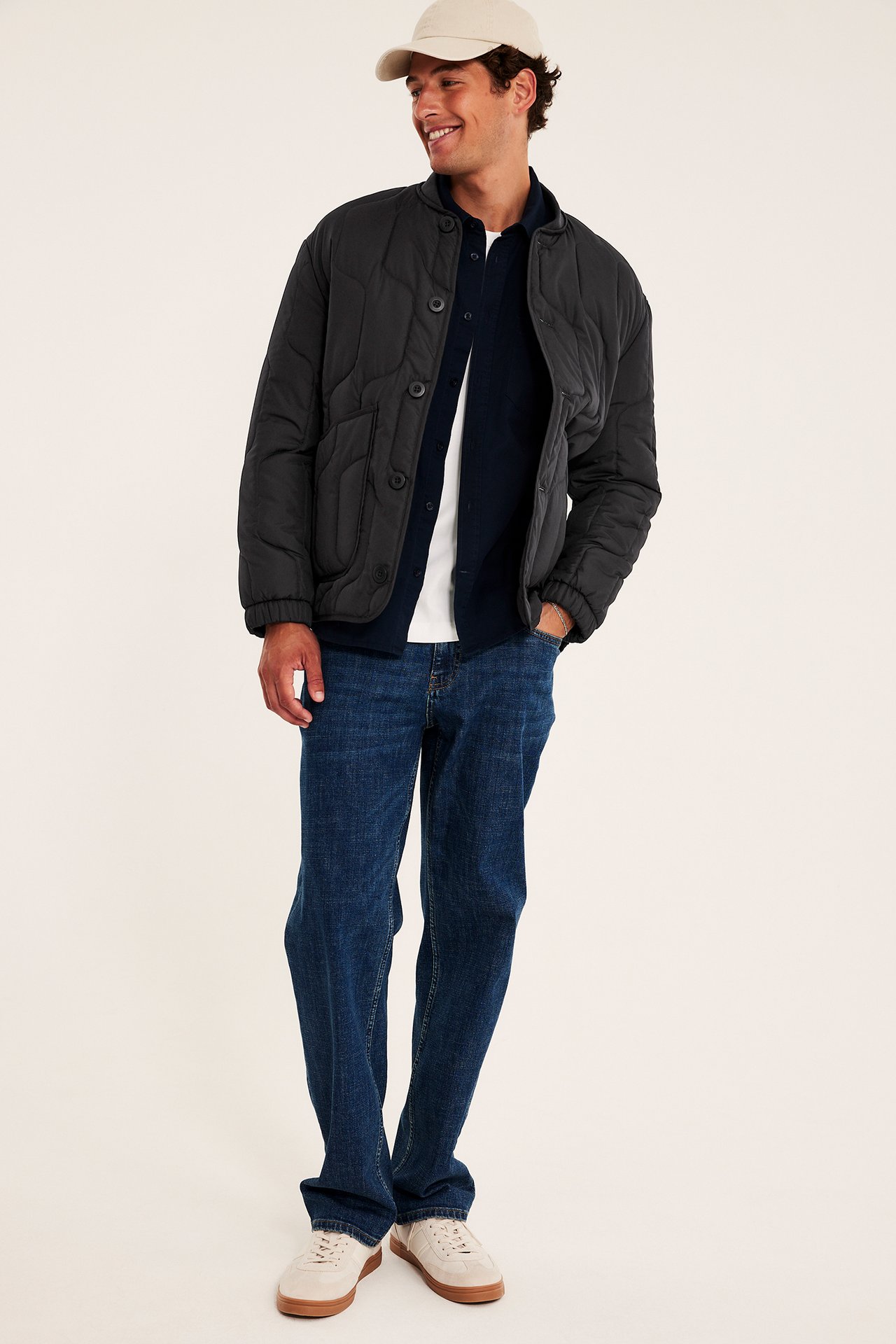 Forfra av mann i mørkegrå vattert jakke, blå jeans, beige caps og joggesko.