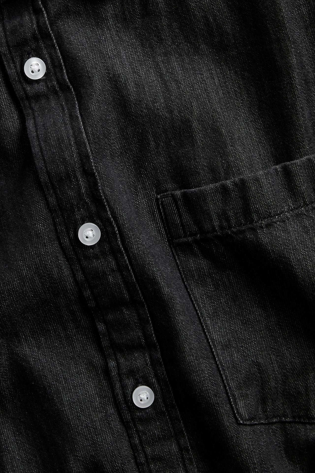 Närbild av en svart denimskjorta med vita knappar och en bröstficka.