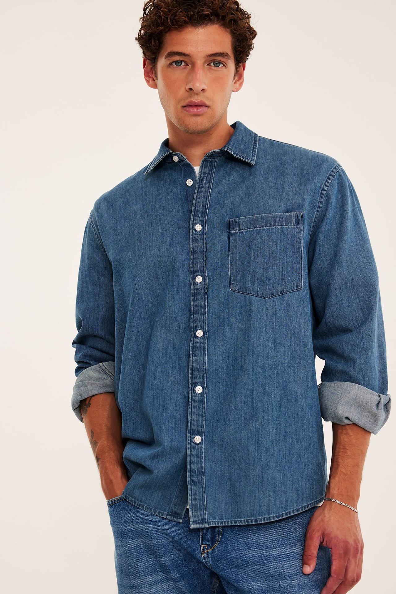 Vuxen man bär blå jeansskjorta, blå jeans och silverkedjearmband, framifrån.