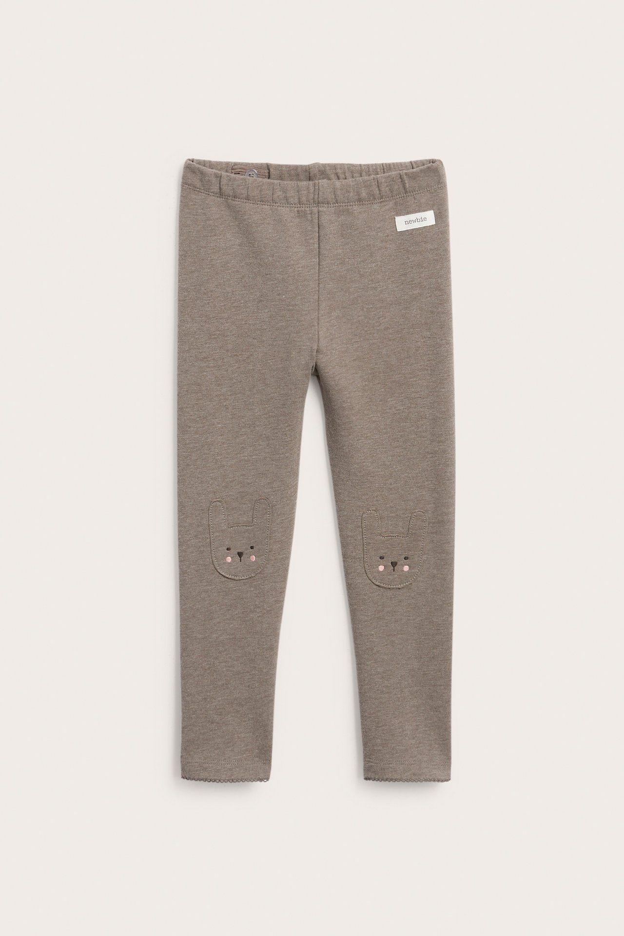 Forfra, taupe baby leggings med kaninøre-lapper på knærne og en 'newbie' etikett.