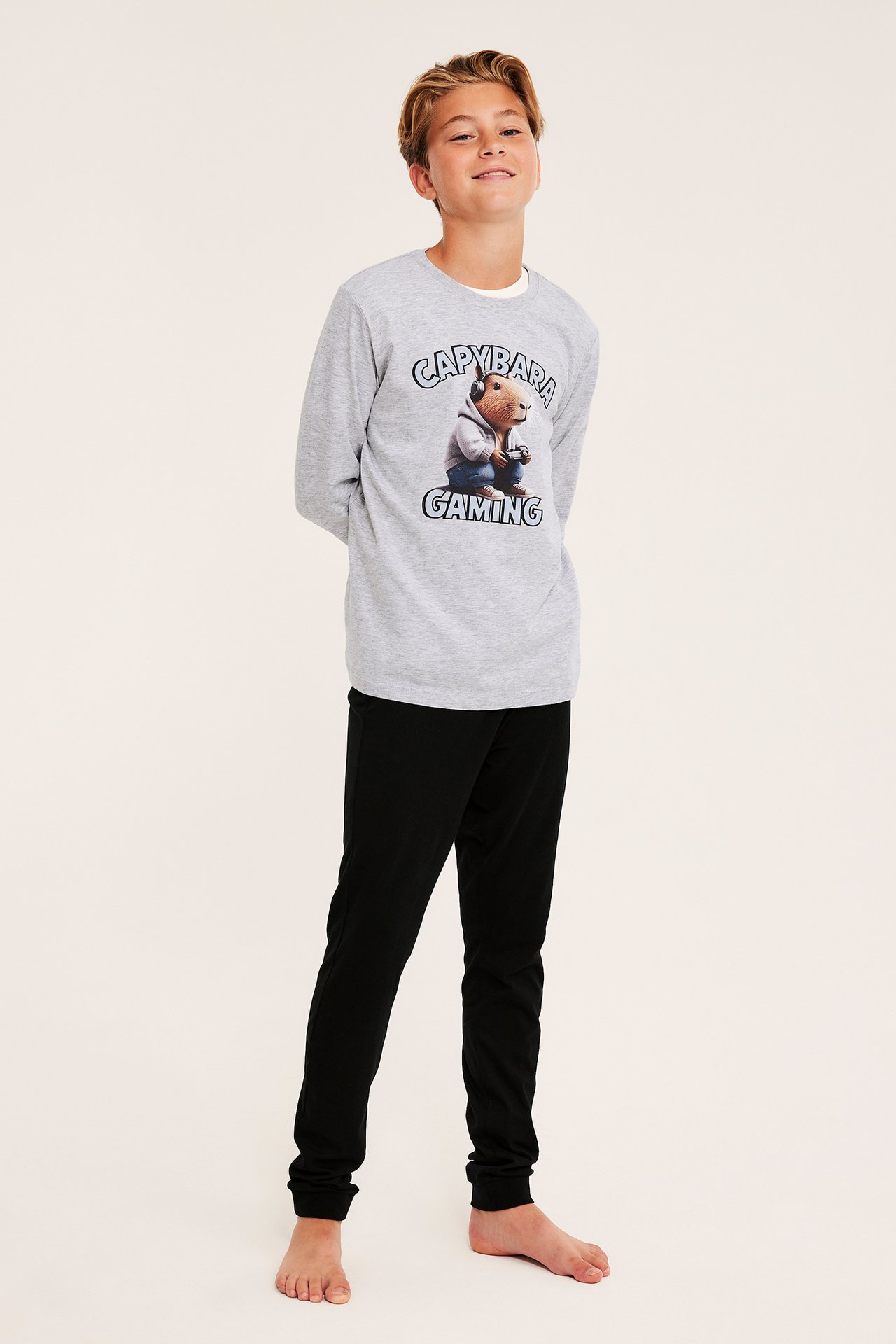 Frontvy av ett barn i grå långärmad t-shirt med capybaratryck och svarta mjukisbyxor.