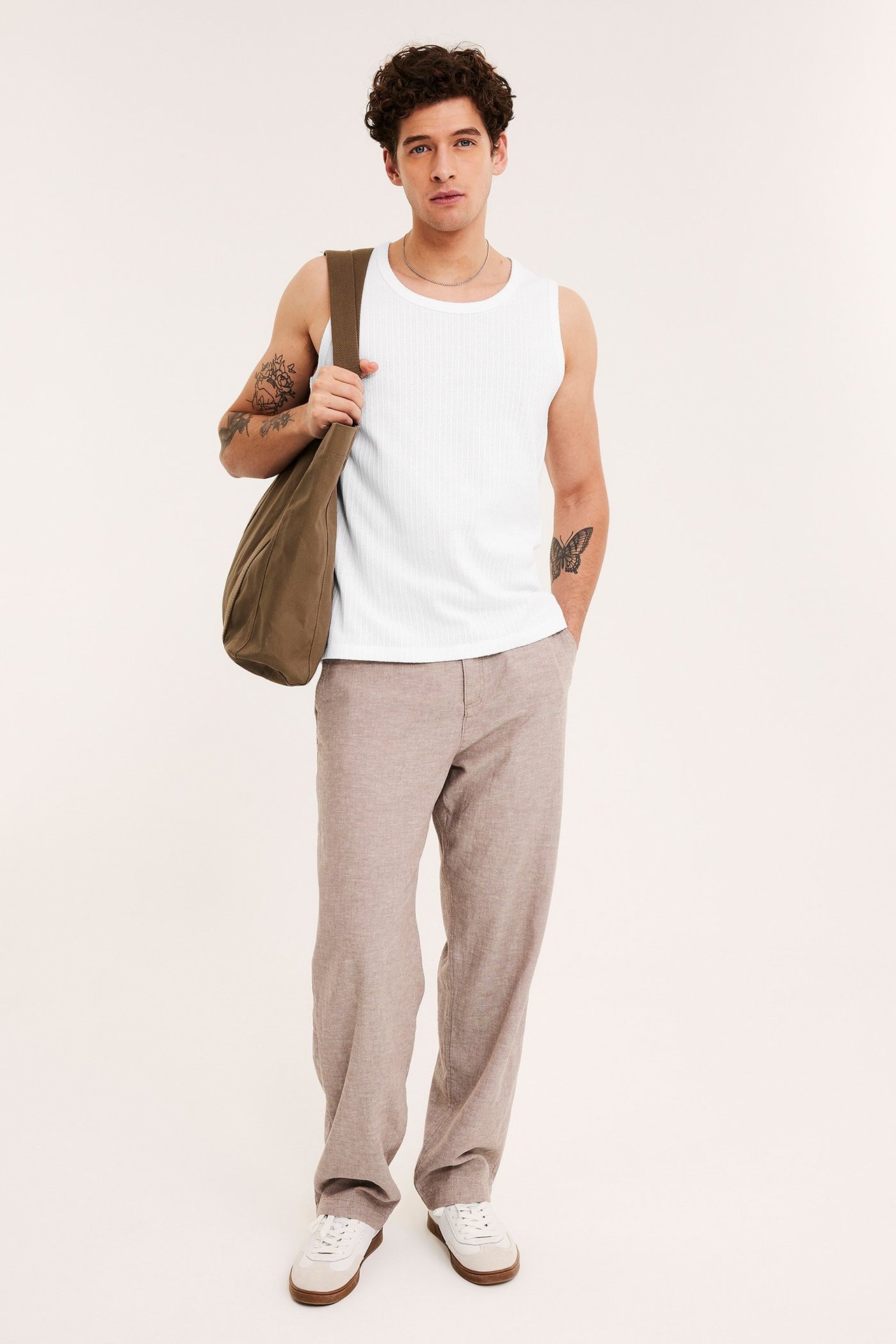 Framifrån: Man bär vitt ribbat linne, ljusbruna byxor, vita sneakers och håller en brun axelväska.