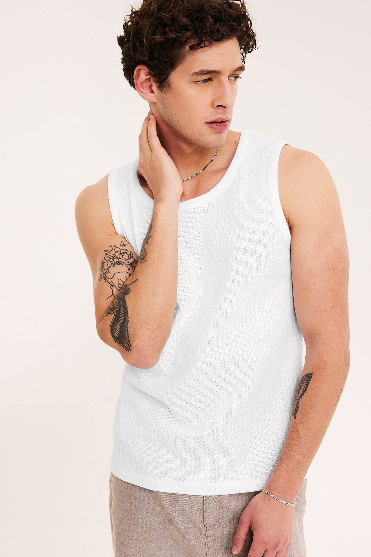 Frontvy av en man i ett vitt pointelle slim fit linne, beige shorts, silverhalsband och armband.