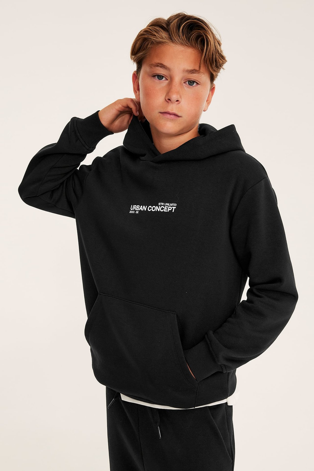 Frontvy av en pojke i en svart hoodie med texttryck och svarta mjukisbyxor.
