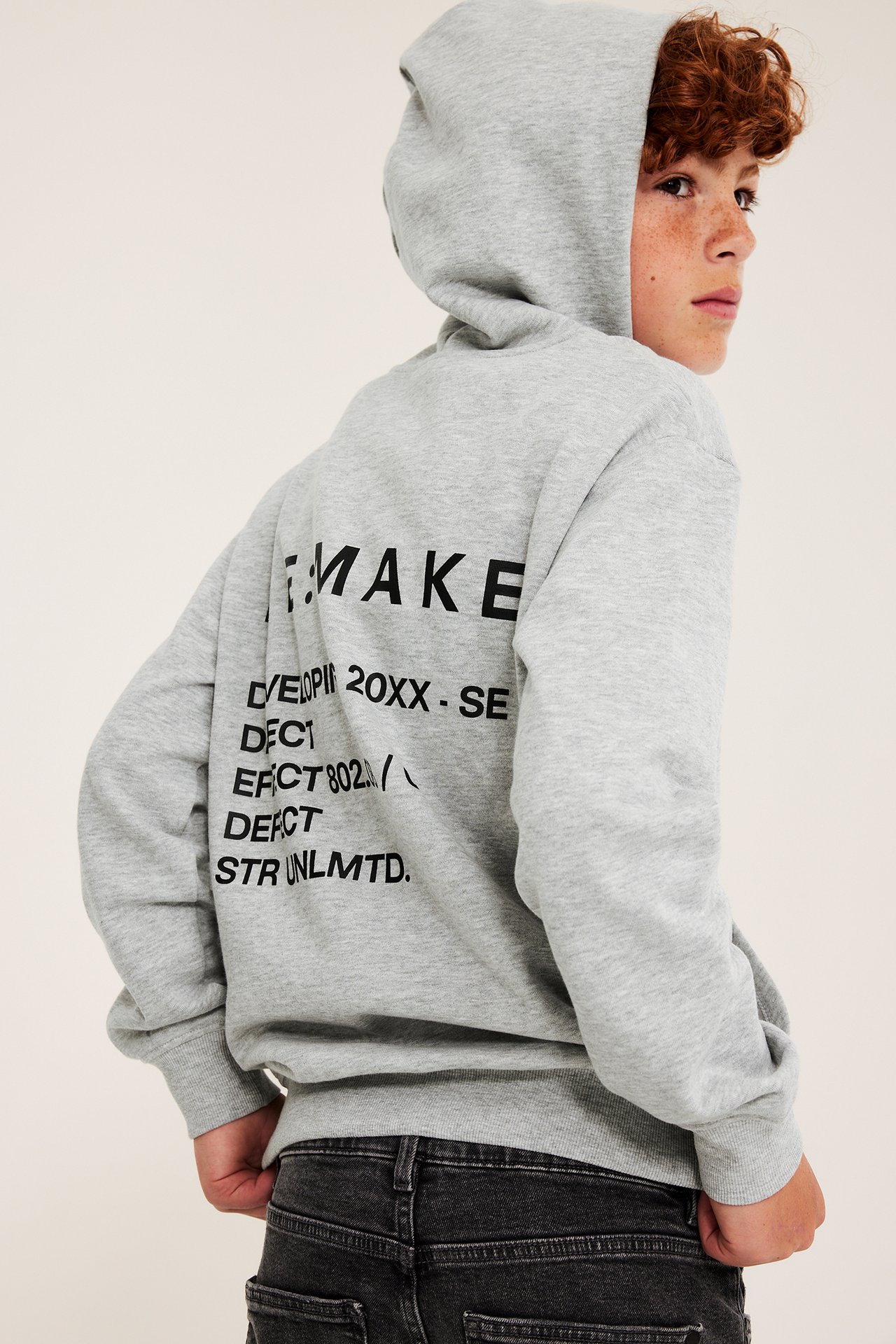 Barns gråa huvtröja med avslappnad passform, svart texttryck på ryggen och svarta jeans, bakifrån.