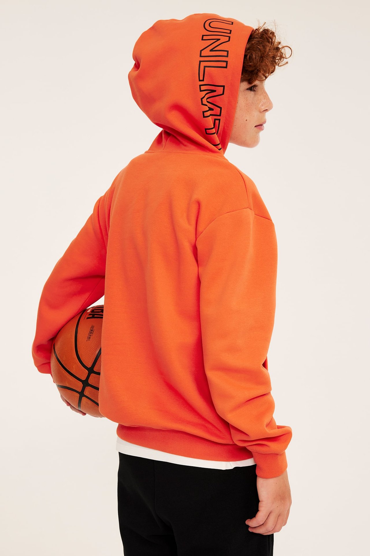 Sidovy av ett barn i en orange huvtröja med UNLMT-tryck, svarta mjukisbyxor, håller en basketboll.