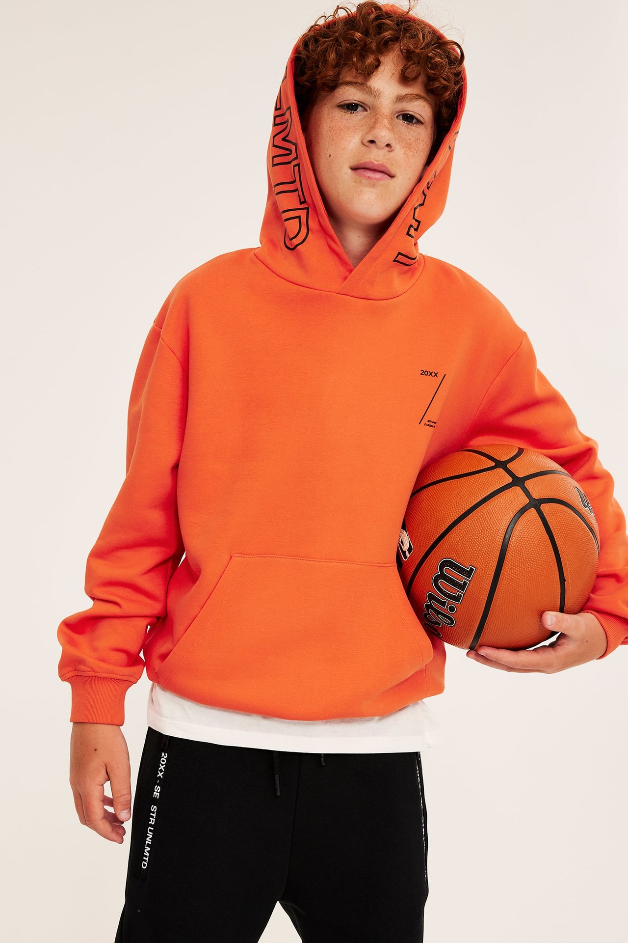 Pojke framifrån i orange huvtröja med svart text, svarta mjukisbyxor och håller i en basketboll.