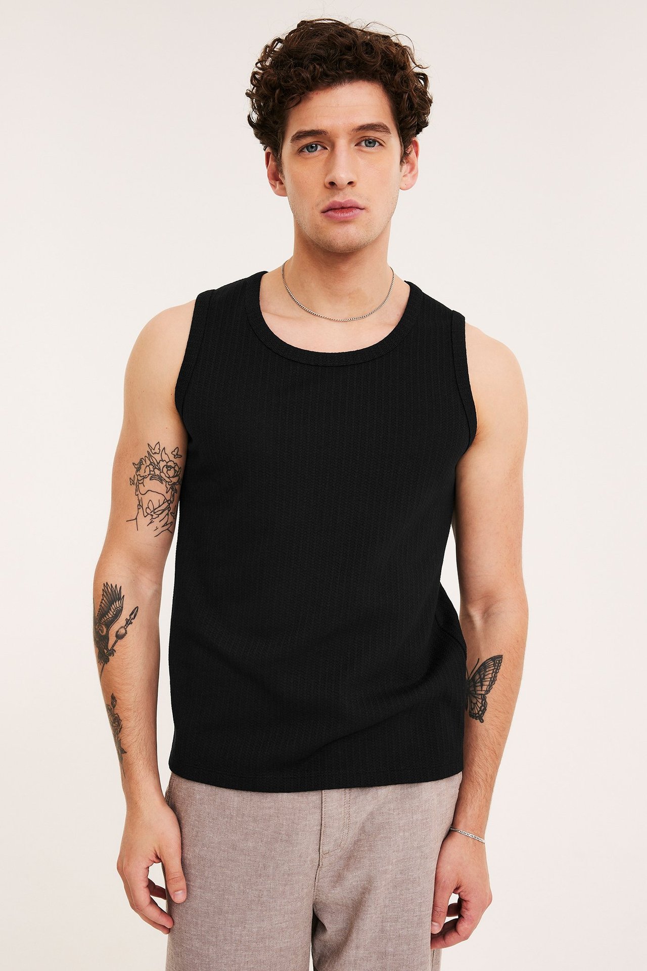 Mann forfra i en svart pointelle-tanktopp, beige shorts og sølvfargede smykker.