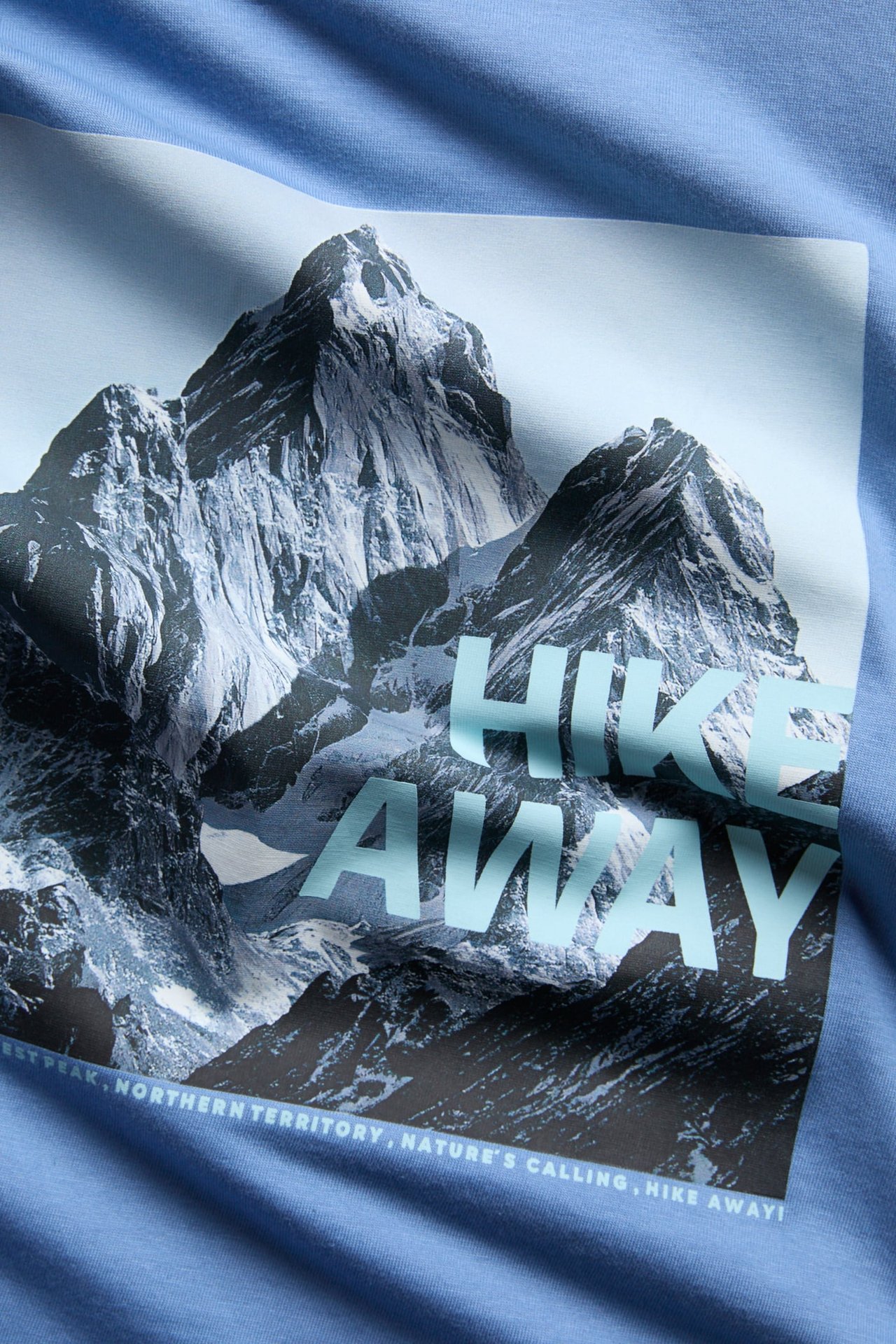 Lähikuva vaaleansinisestä t-paidasta, jossa on vuoristokuvio ja teksti 'HIKE AWAY'.