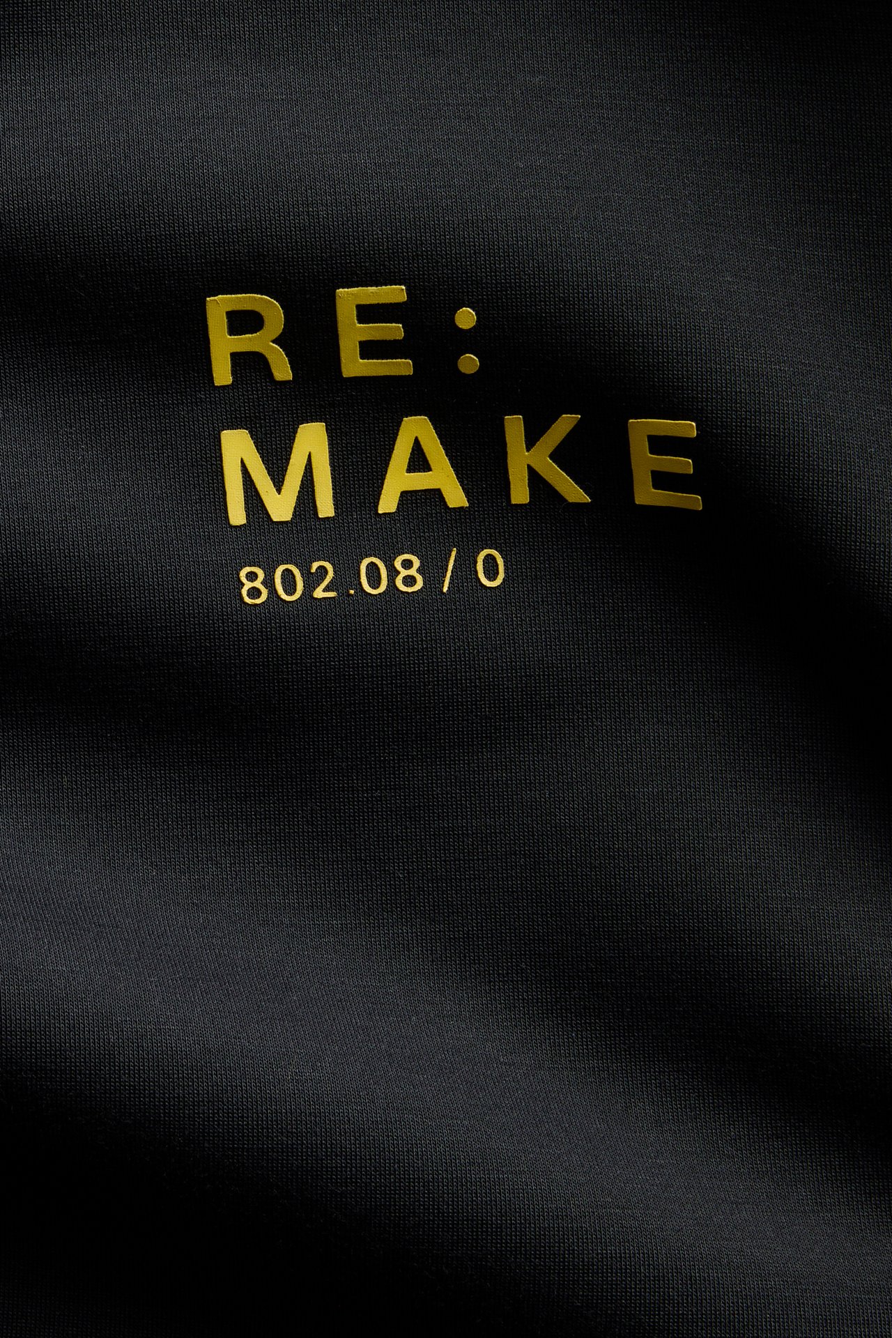 Närbild på ett svart plagg med guldtexten 'RE: MAKE' och numren '802.08 / 0'.
