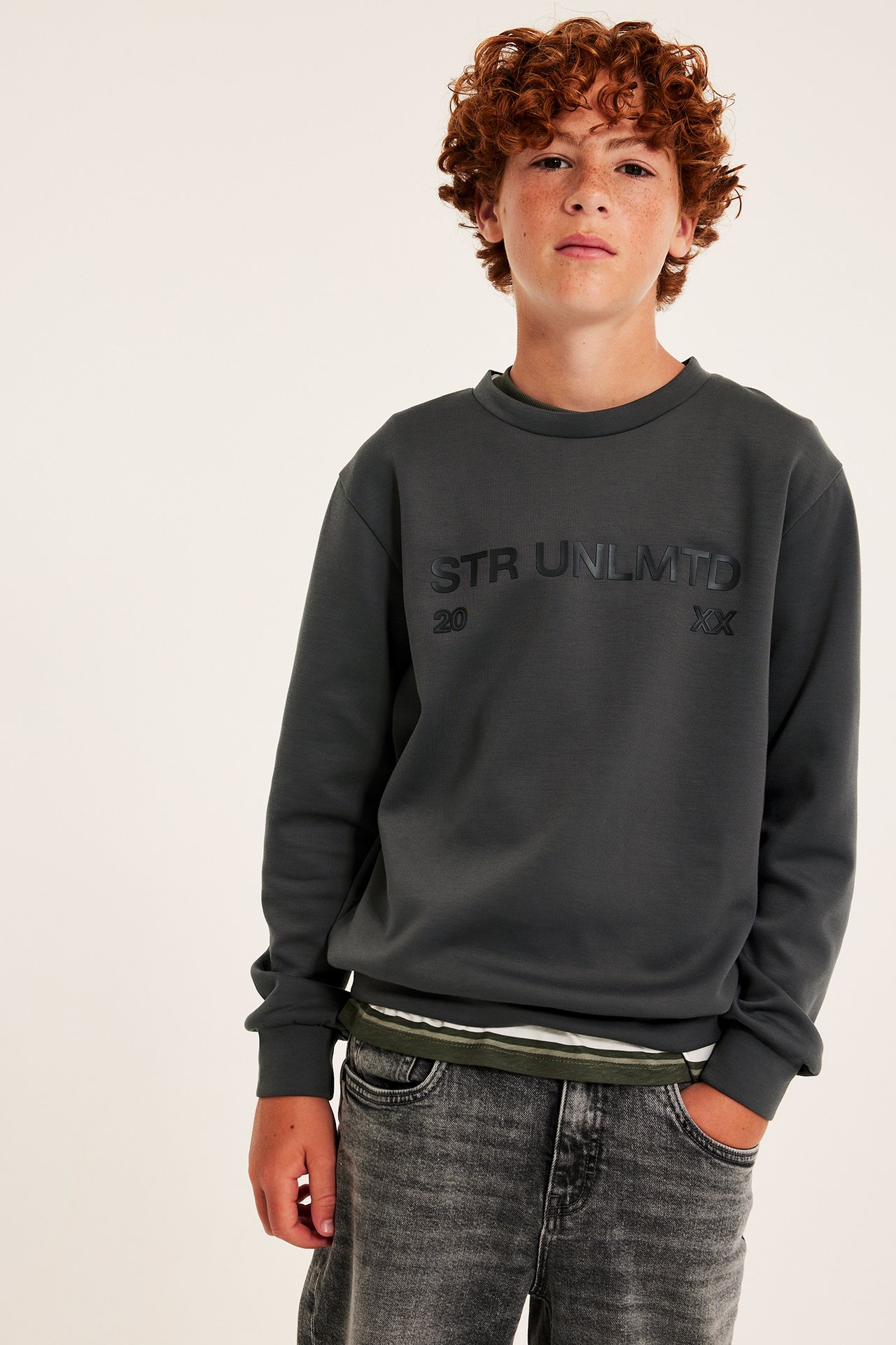 Framsida: Barnens mörkgrå sweatshirt med texttryck, bärs över en randig t-shirt och svarta jeans.