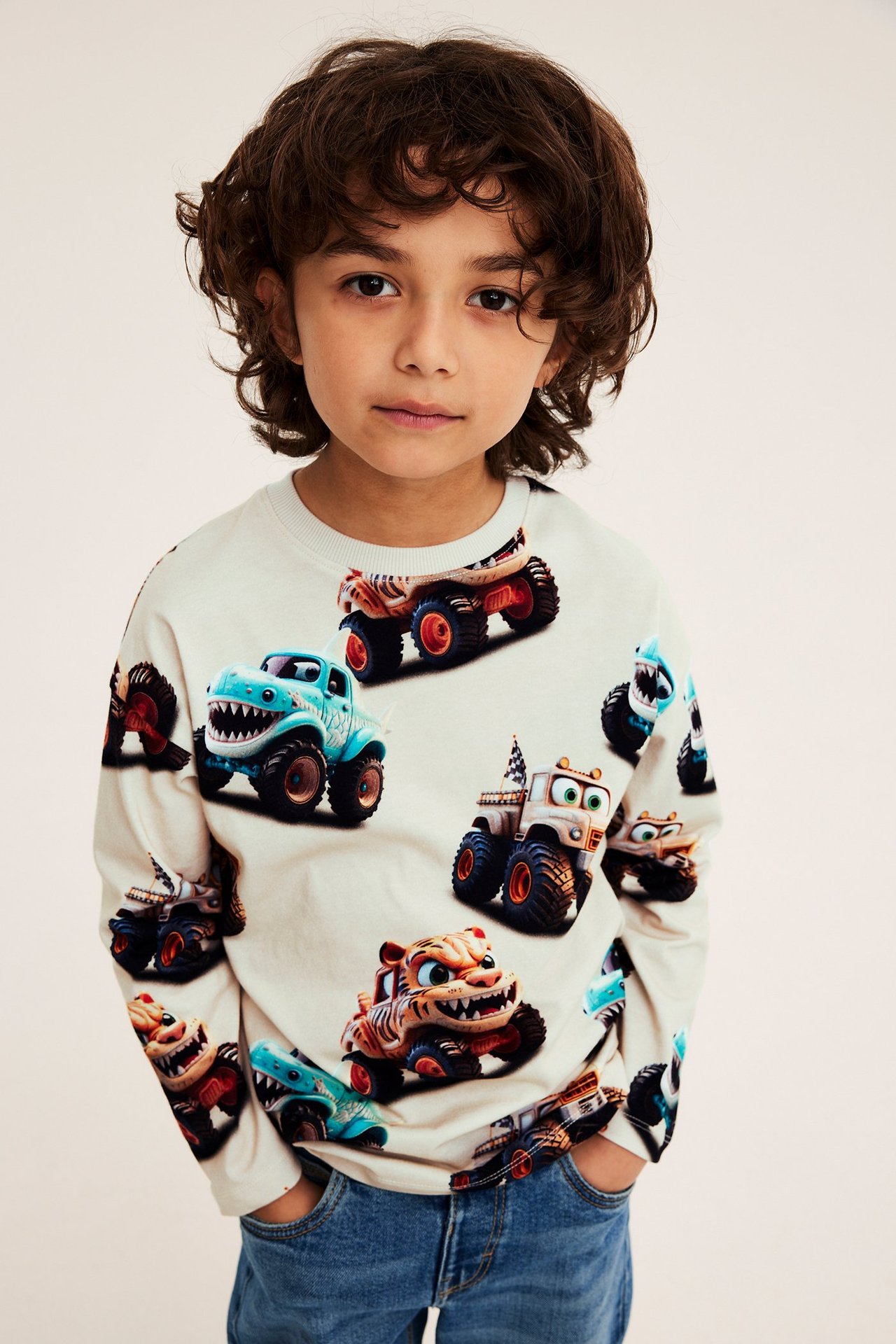 Framifrån: ett barn i ljusbeige långärmad t-shirt med monstertruckprint och blå jeans.