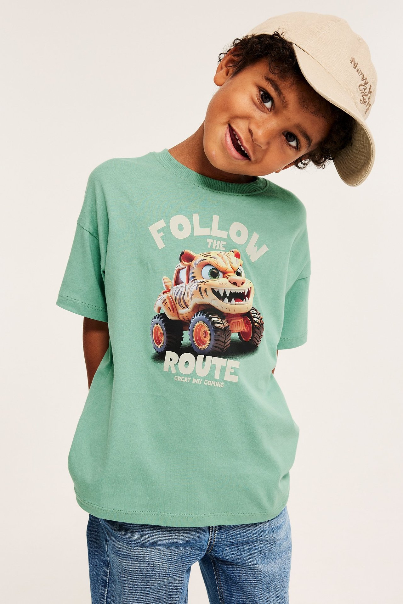 Barn bär grön bomulls-T-shirt med tigermonstertruck-tryck, blåa jeans och en beige keps.