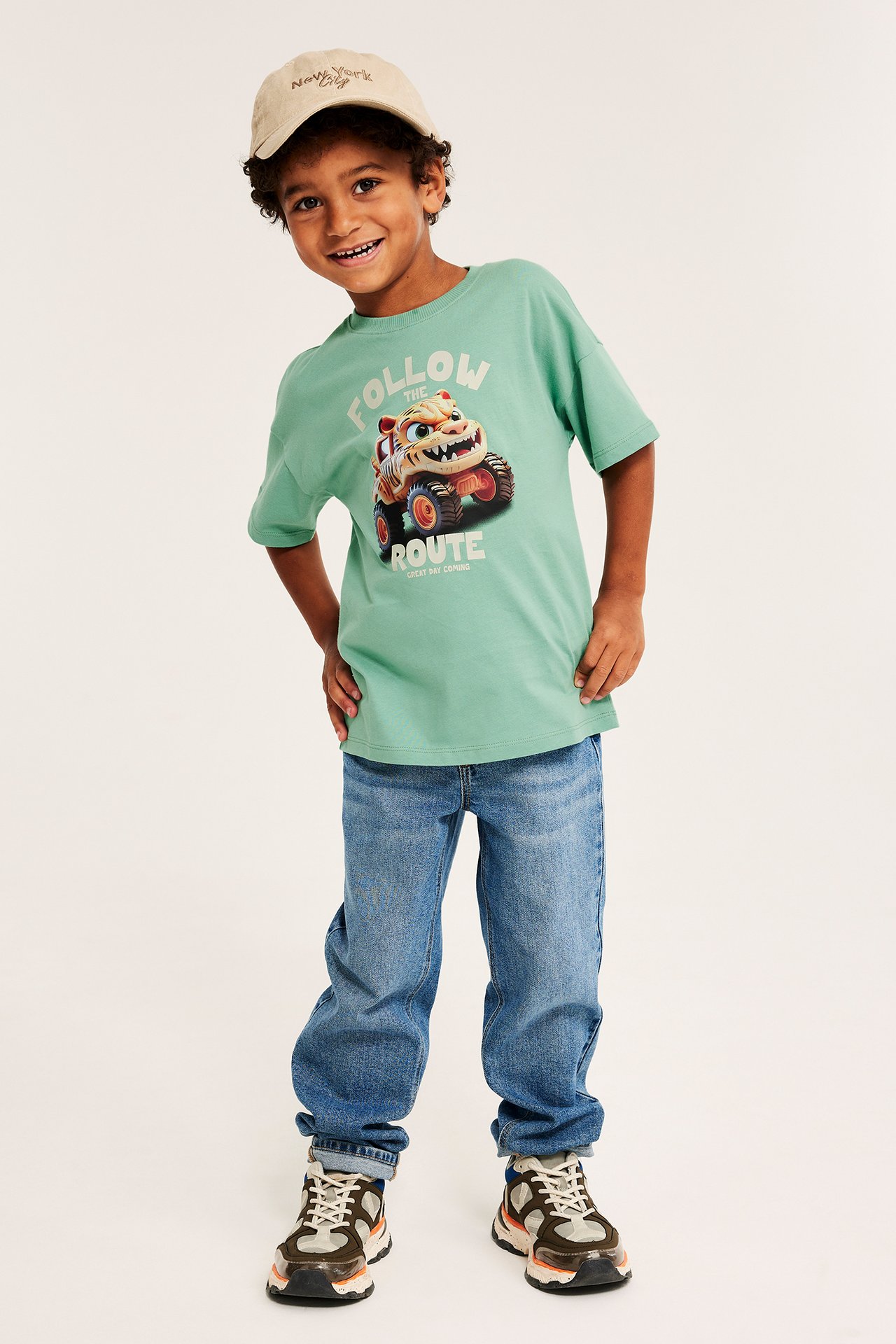 Framifrån, ett barn i beige keps, grön T-shirt med tigermonstertruck-tryck, blå jeans och sneakers.