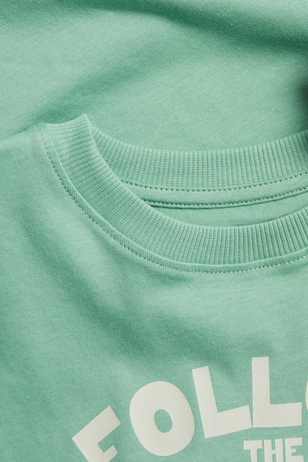 Zbliżenie zielonej bawełnianej koszulki dziecięcej z prążkowanym dekoltem i białym nadrukiem tekstowym z przodu.