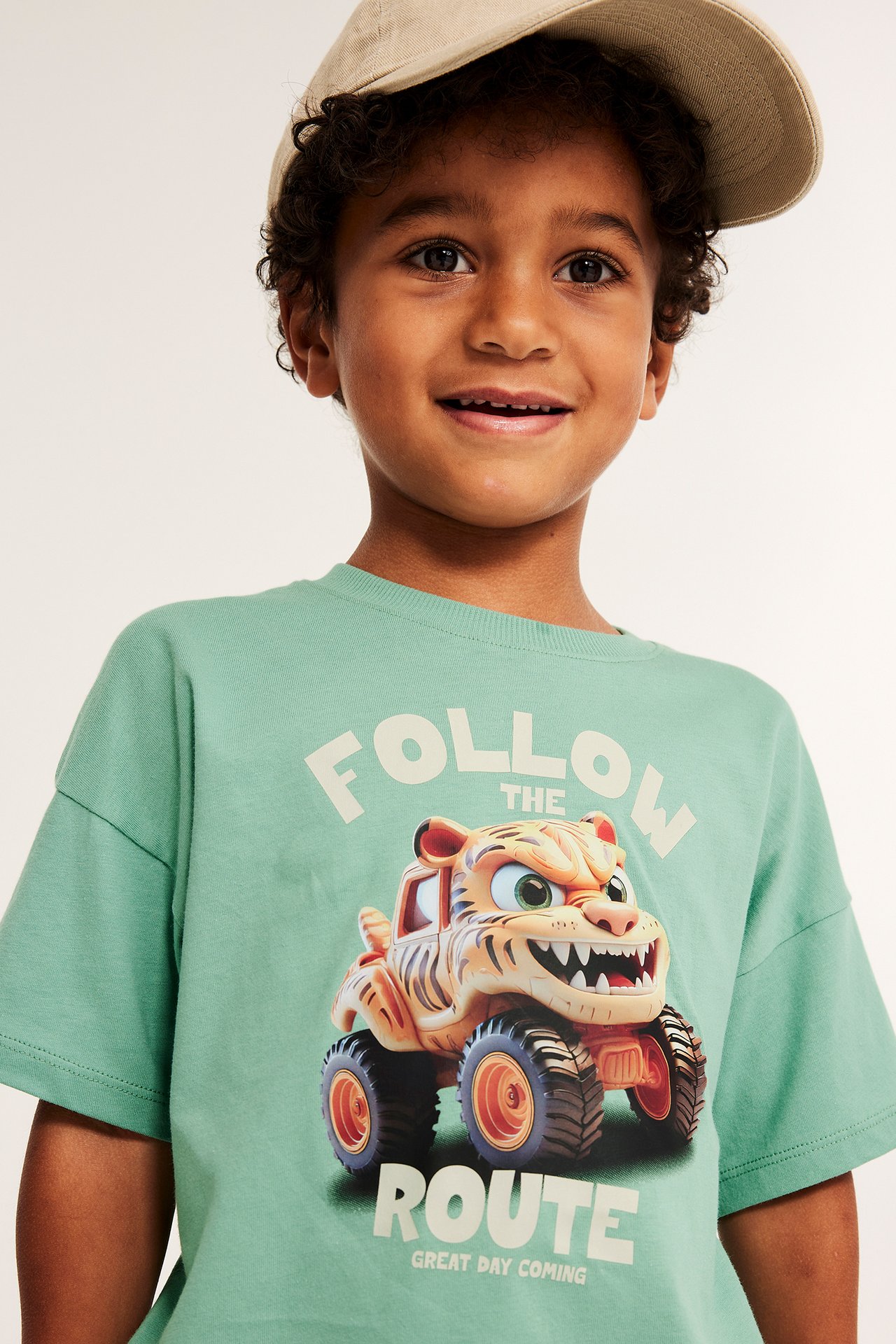 Framifrån: Barn bär grön bomulls-t-shirt med tryck av tigermonstertruck och beige keps.