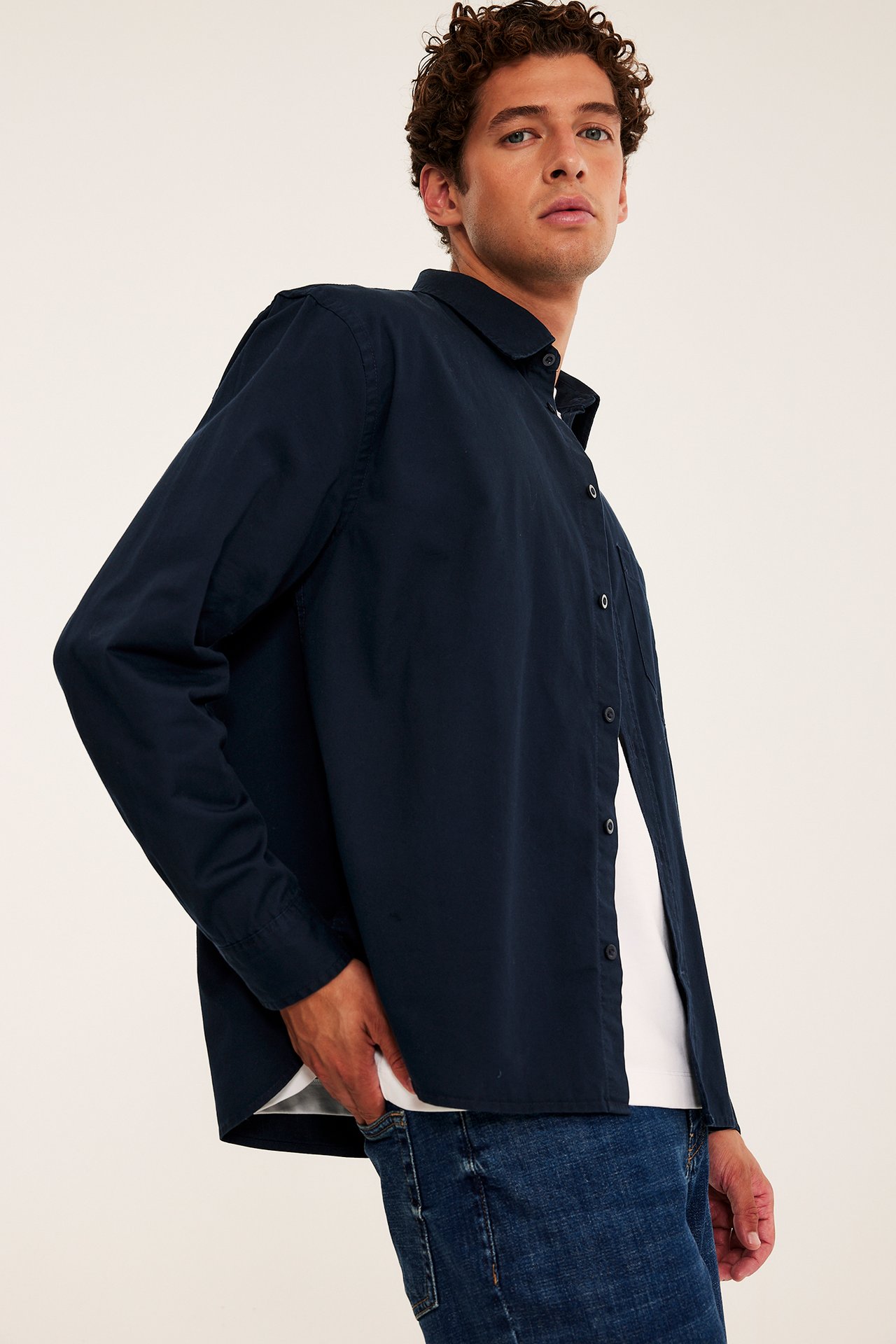 Voksen mann i sidestilling med mørkeblå langermet skjorte over hvit t-skjorte og blå jeans.
