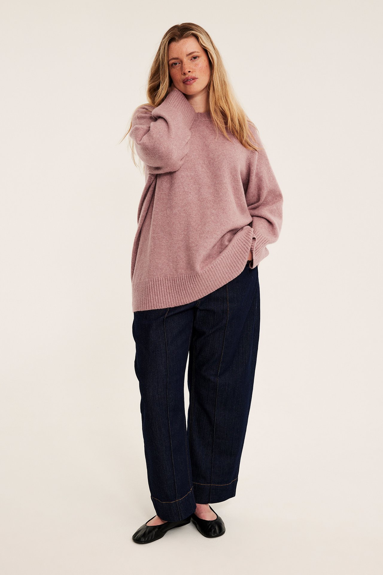 Framifrån, kvinnlig modell i lös rosa stickad tröja, mörkblå vida jeans och svarta ballerinaskor.