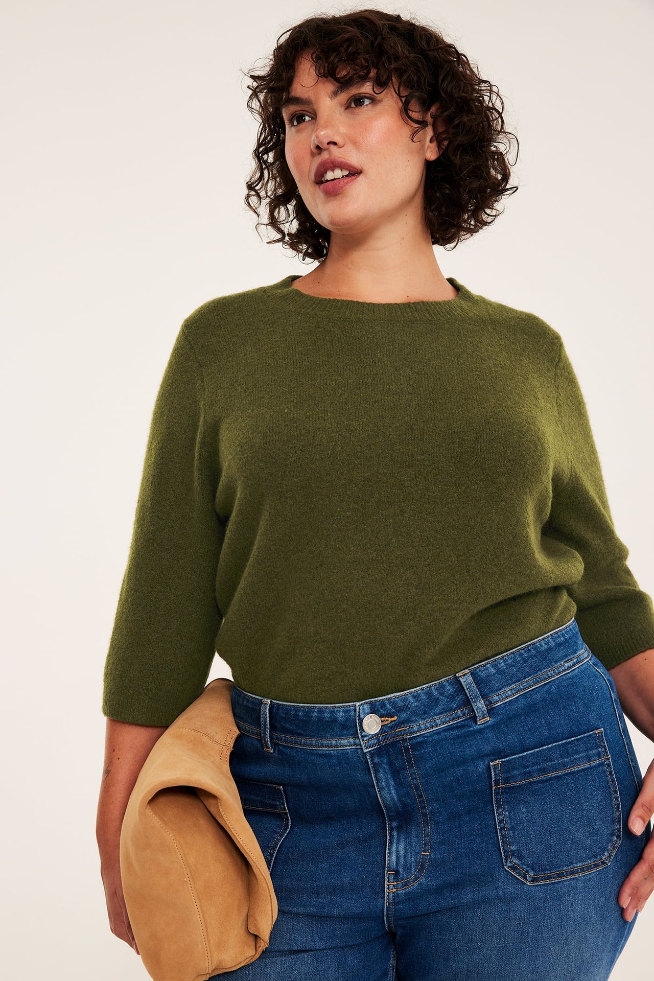 Frontvy: Kvinnlig modell i olivgrön tröja, blå jeans, håller en beige mockaväska.