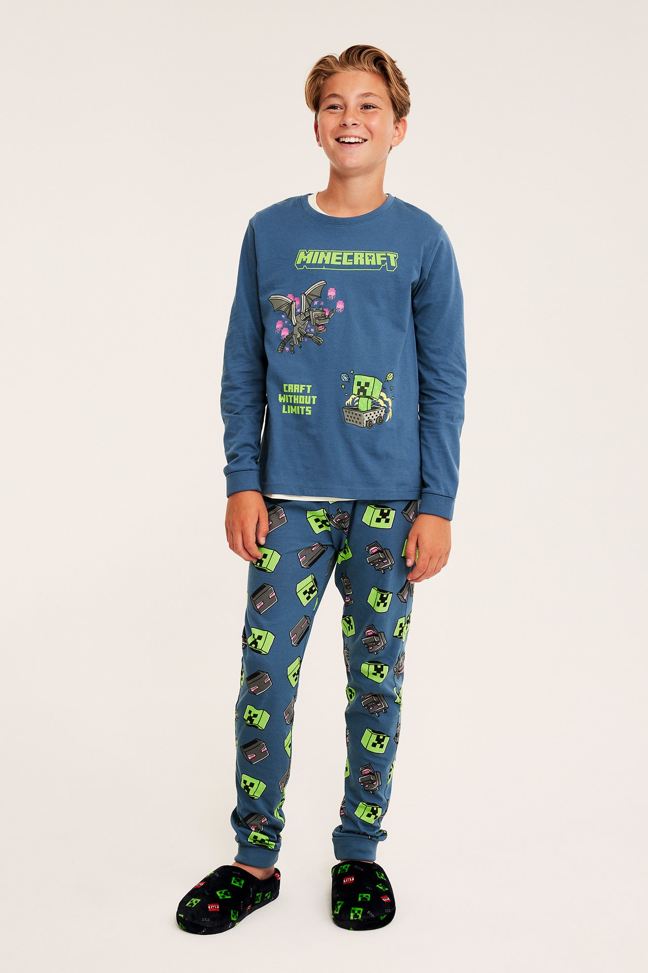 Edestä: poika petrolinsinisessä Minecraft-kuvioisessa pitkähihaisessa pyjamapaidassa, yhteensopivissa housuissa ja mustissa tohveleissa.