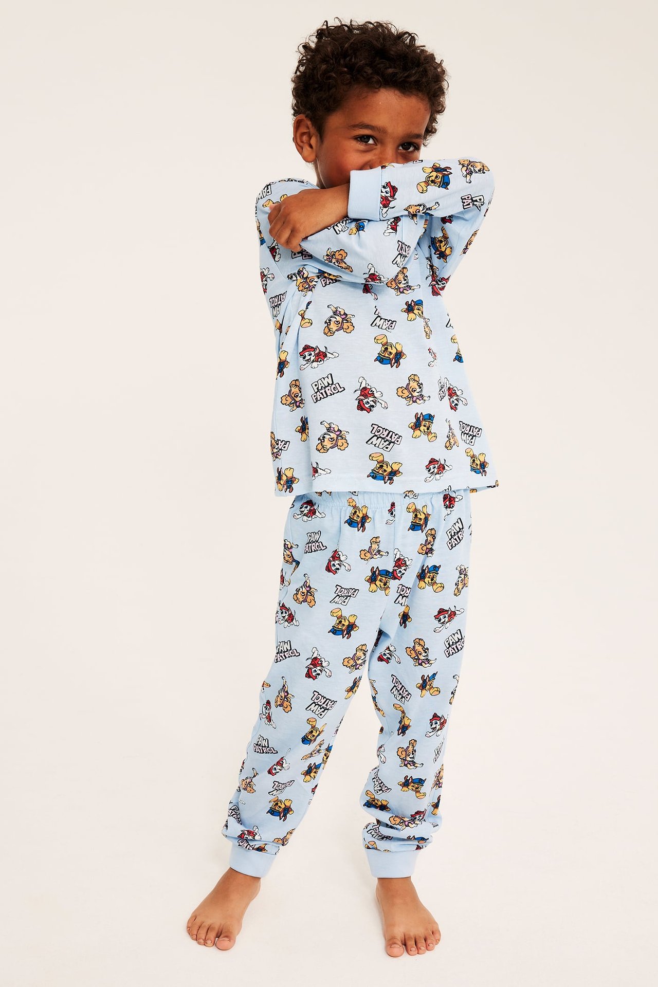 Edestä näkymä: Lapsi yllään vaaleansininen Paw Patrol -kuvioinen pyjama, jossa resorit ranteissa ja nilkoissa.