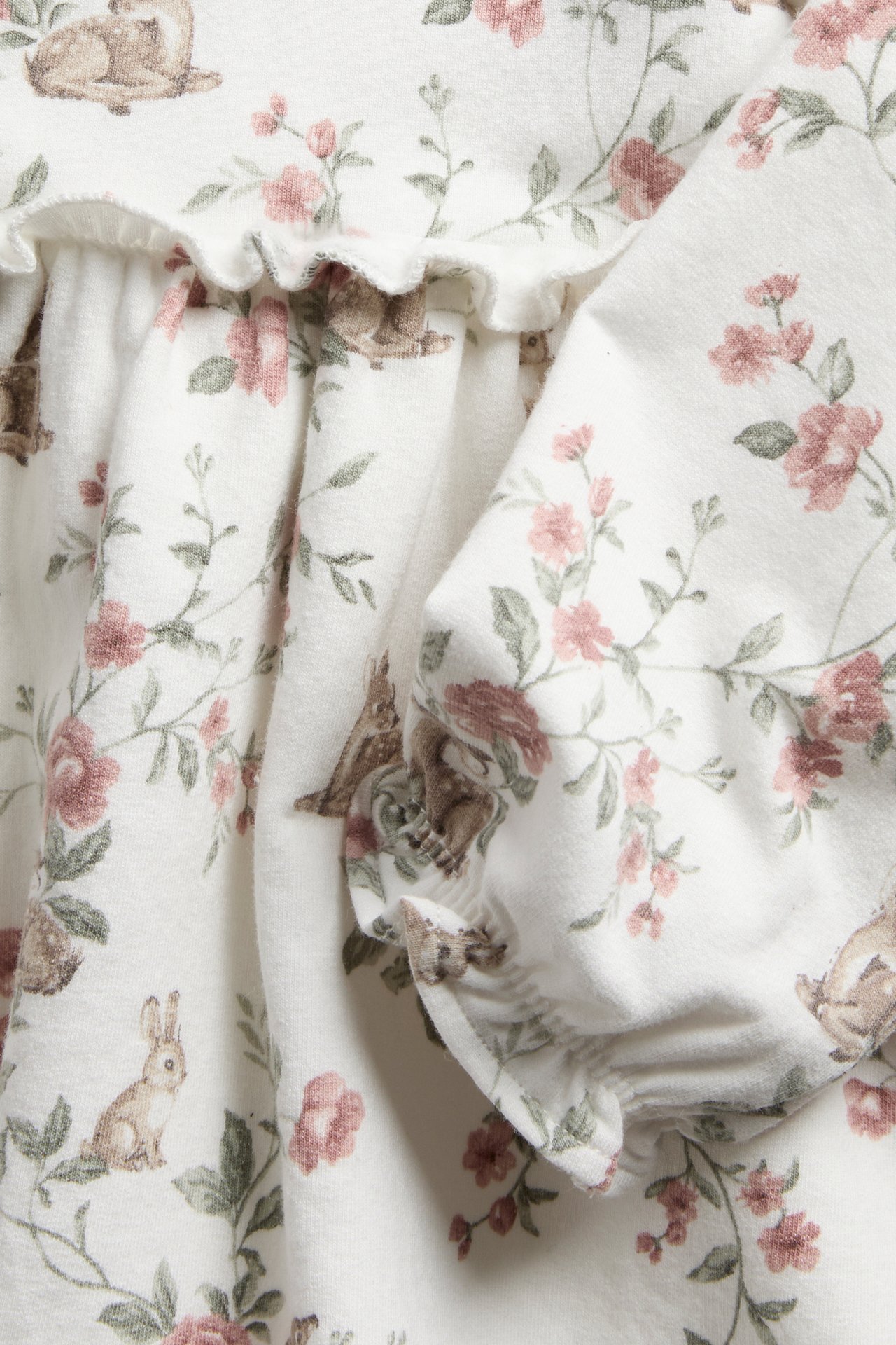 Närbild av en offwhite jersey barnklänning med brunt kanin- och rosa blomstermönster, med volangdetalj.