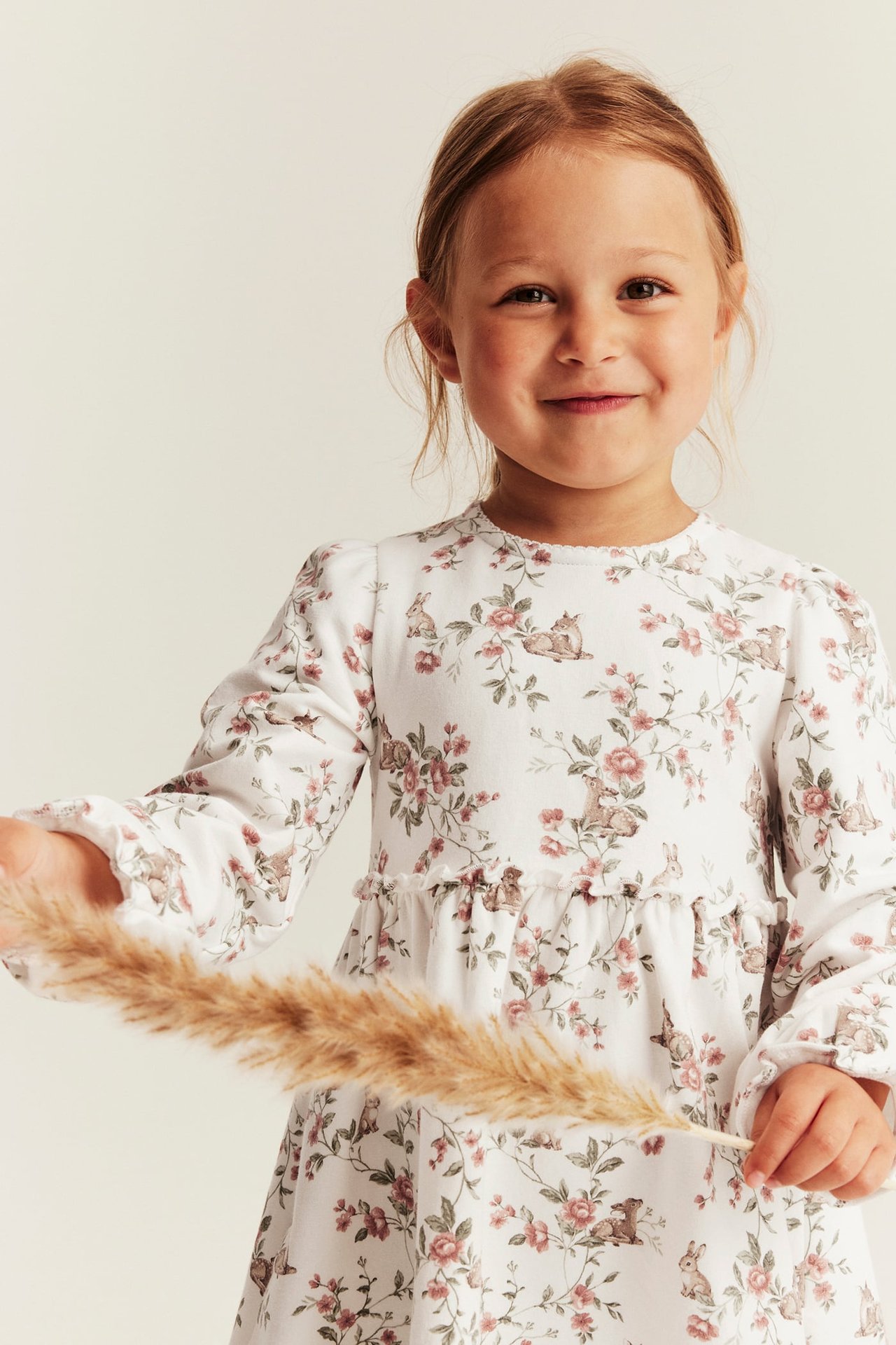 Frontutsikt av et barn i offwhite jerseykjole med blomster- og kaninmønster, holder en tørket plante.