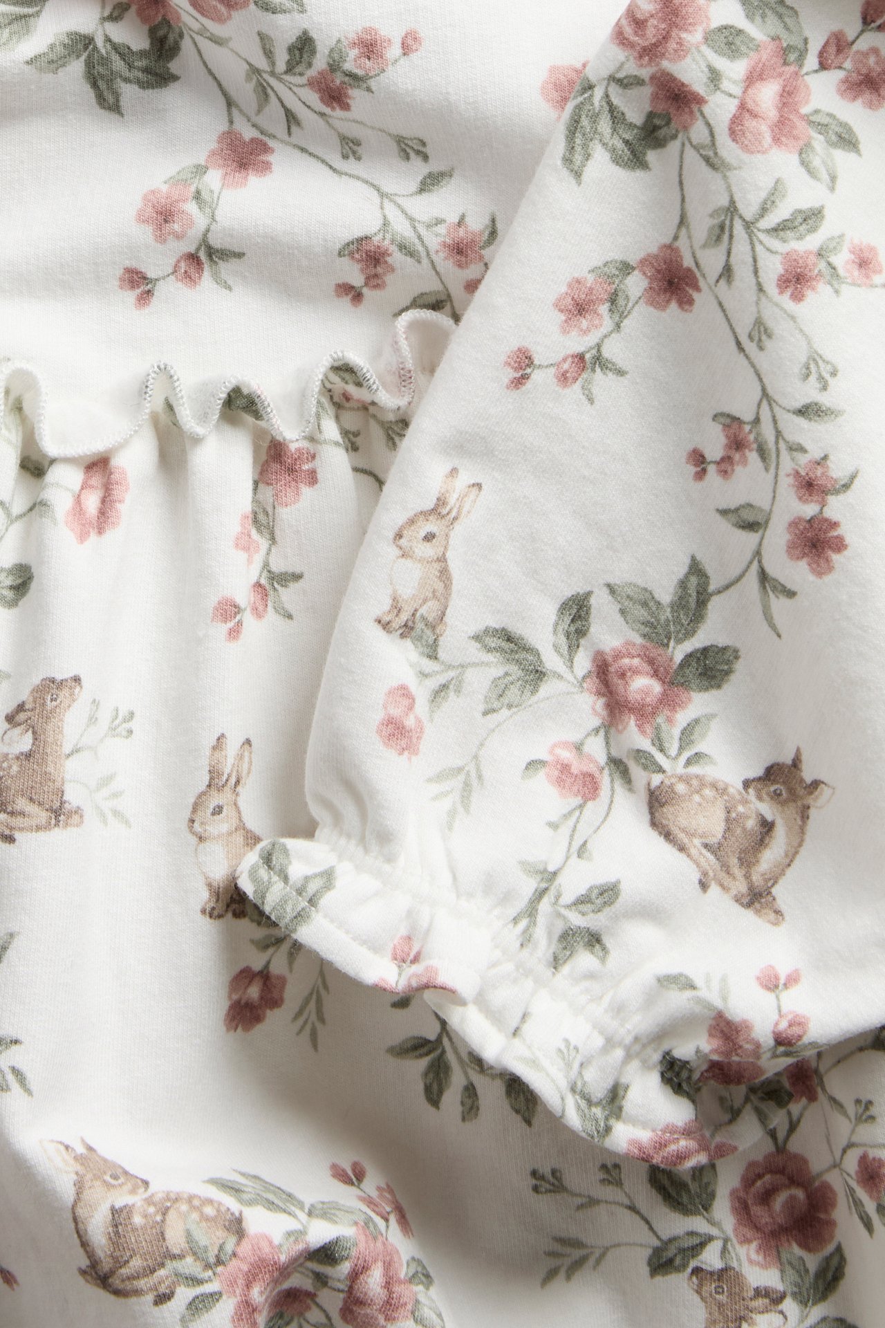 Närbild av en benvit baby-jerseyklänning med rosa blommor, rådjur och kaniner, samt volangdetaljer.