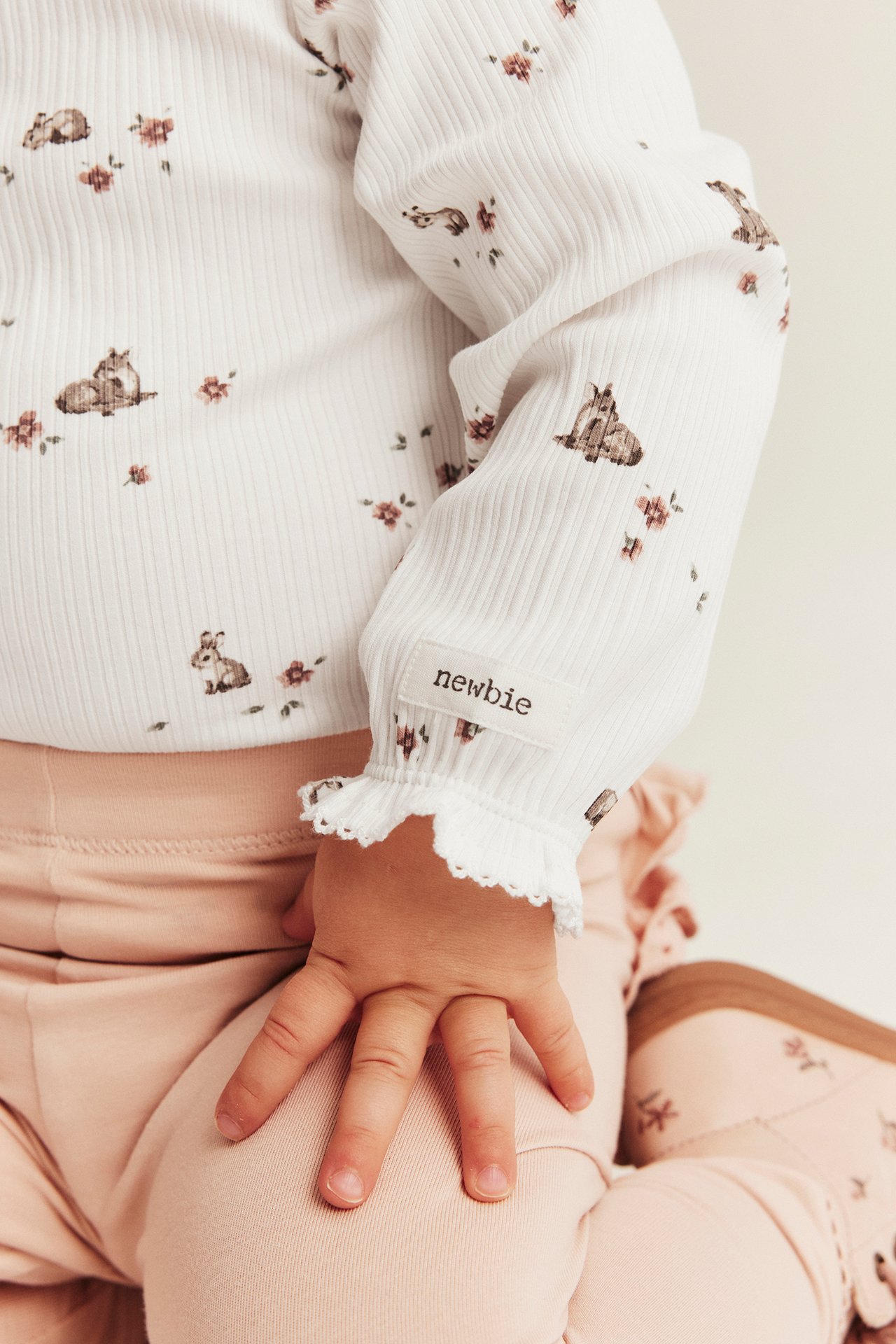 Nærbilde av baby i off-white ribbet body med rådyr- og blomstermønster, volangmansjetter og rosa bukse.
