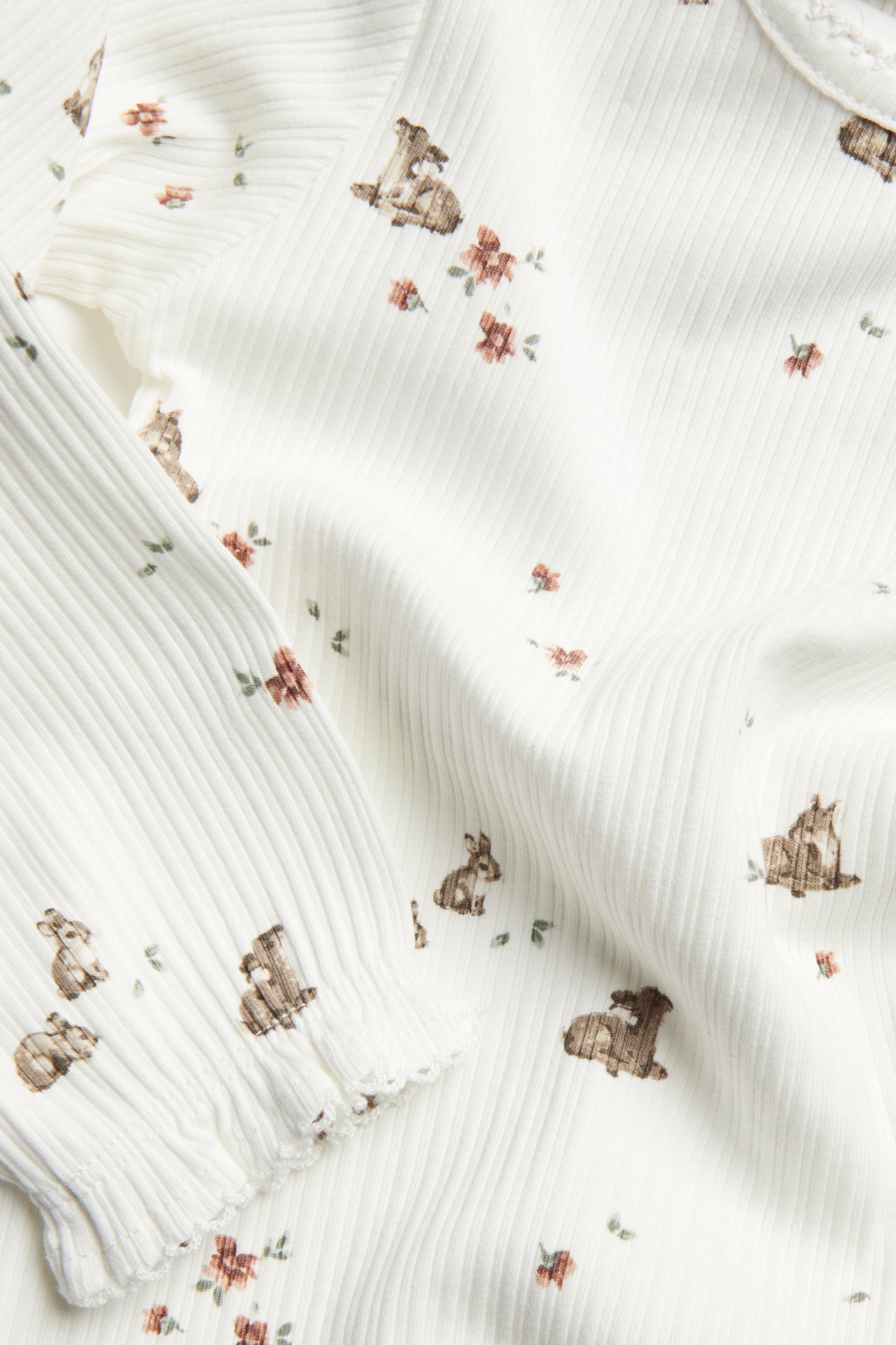 Nærbilde av en offwhite ribbet langermet babybody med dyre- og blomstermønster, med volanger.