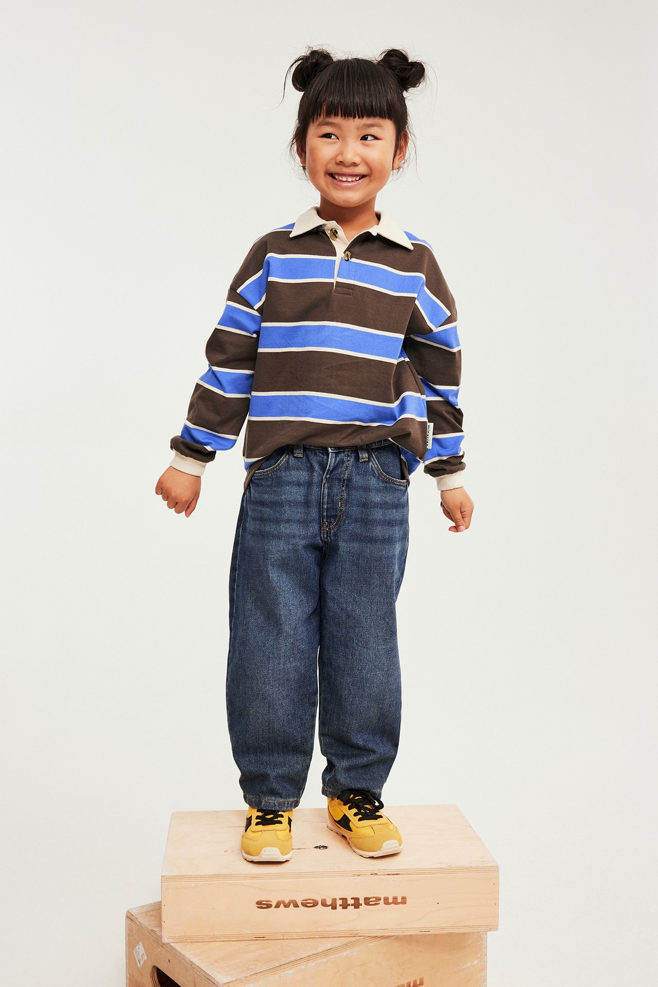 Forfra, barn i brunstripet langermet poloskjorte, blå jeans og gule sneakers.