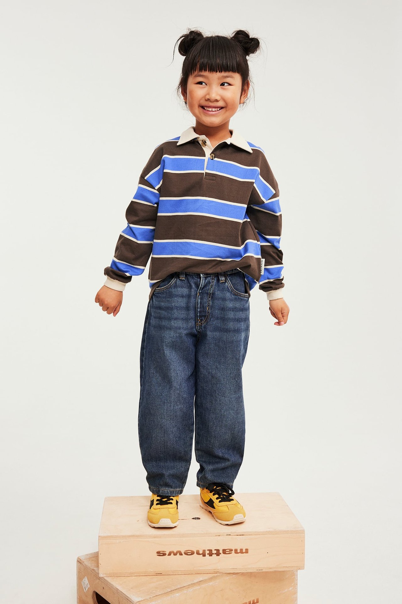 Forfra, barn i brunstripet langermet poloskjorte, blå jeans og gule sneakers.