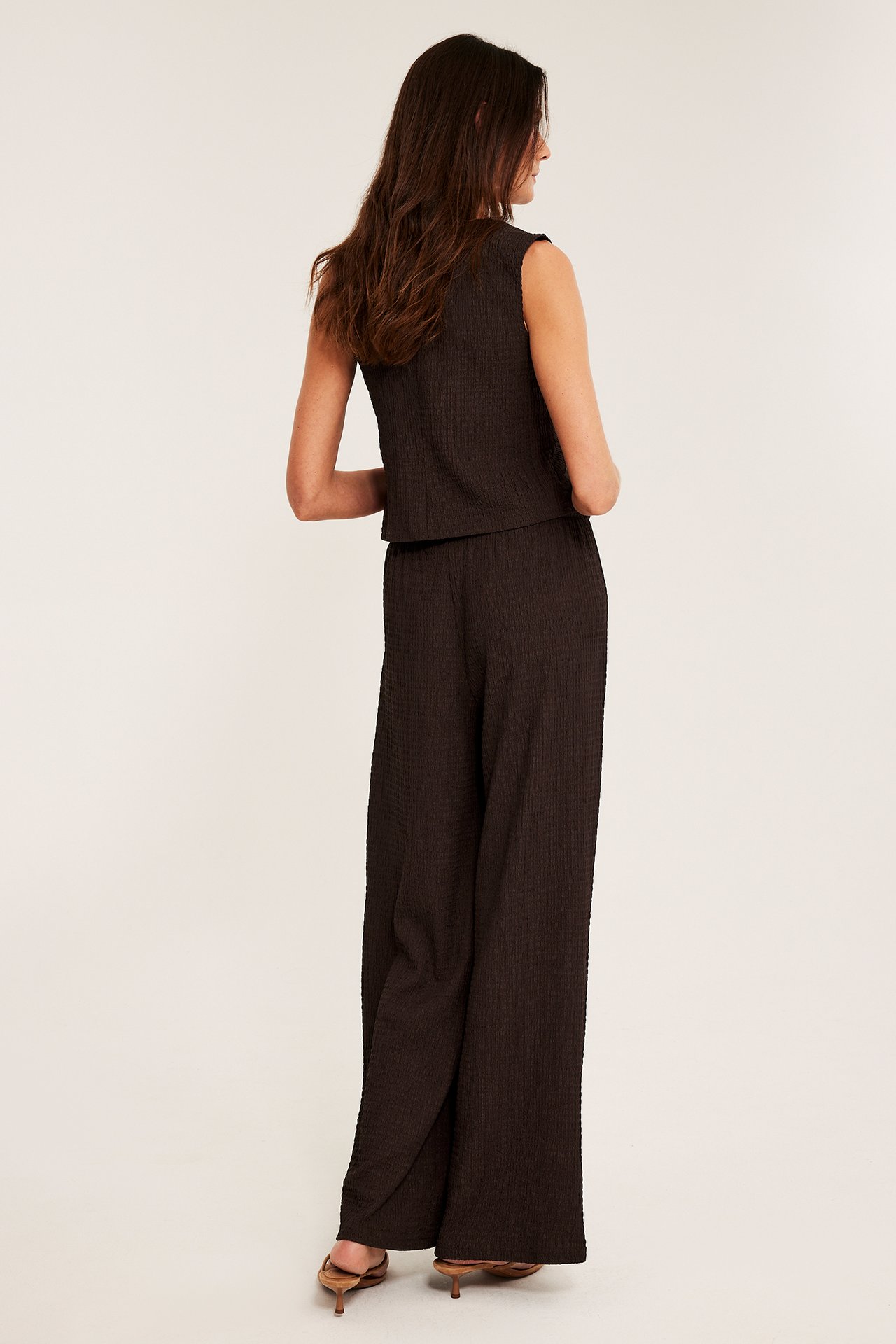 Takaa: nainen tummanruskeissa kuvioiduissa leveälahkeisissa trikoohousuissa, yhteensopiva toppi ja ruskeat korkokengät.