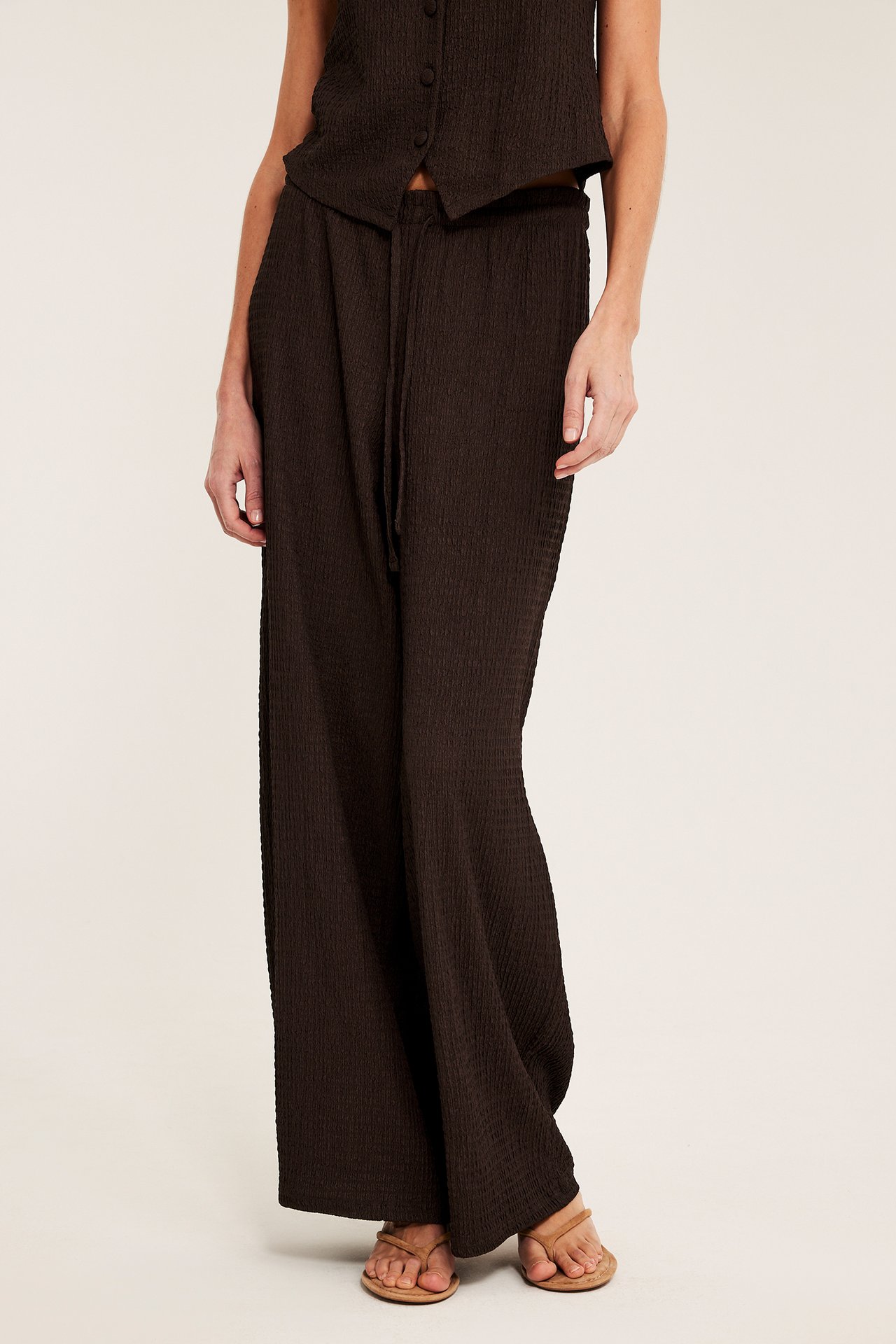 Edestä: Nainen yllään tummanruskeat joustavat housut, yhteensopiva toppi ja vaaleanruskeat sandaalit.