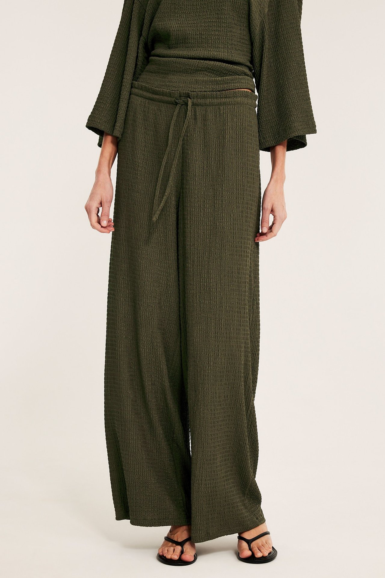 Frontvy: Kvinna bär gröna strukturerade jerseybyxor med dragsko, matchande topp och svarta sandaler.